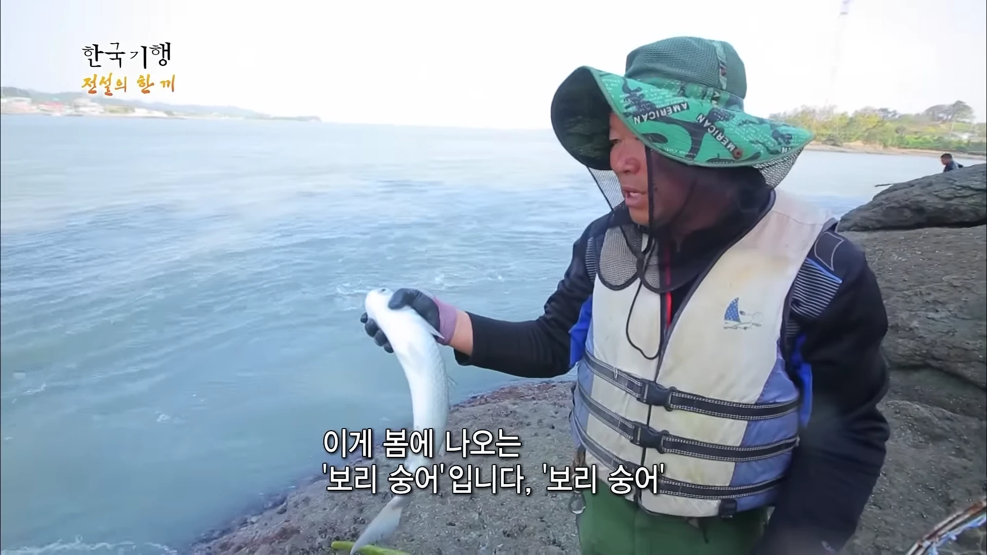바닷가 바위 위에서 챙이 넓은 모자와 구명조끼를 착용한 어부가 직접 잡은 숭어를 손에 들고 설명하고 있다.