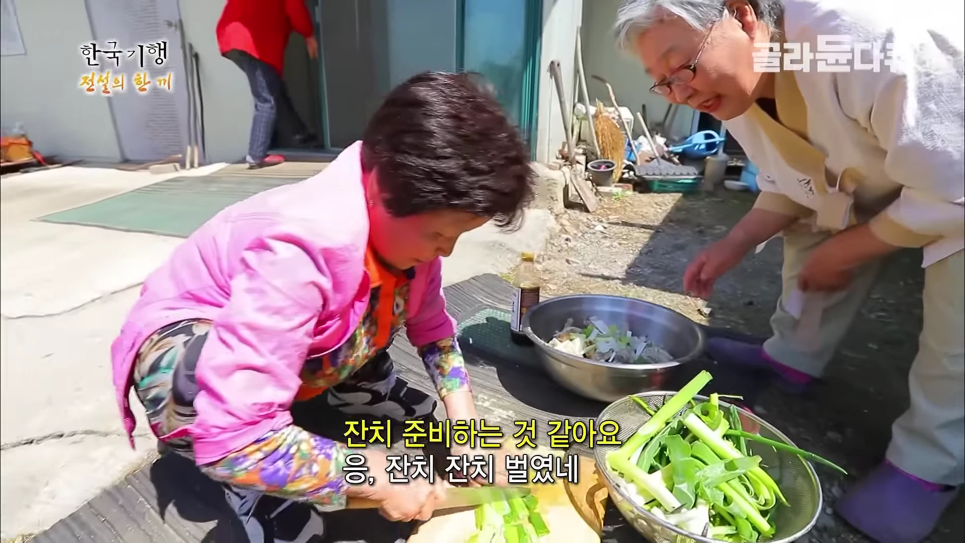 마당에서 한 여성이 쪼그려 앉아 도마 위에서 파를 썰고 있고, 그 옆에 다른 여성이 서서 지켜보고 있는 모습이다.