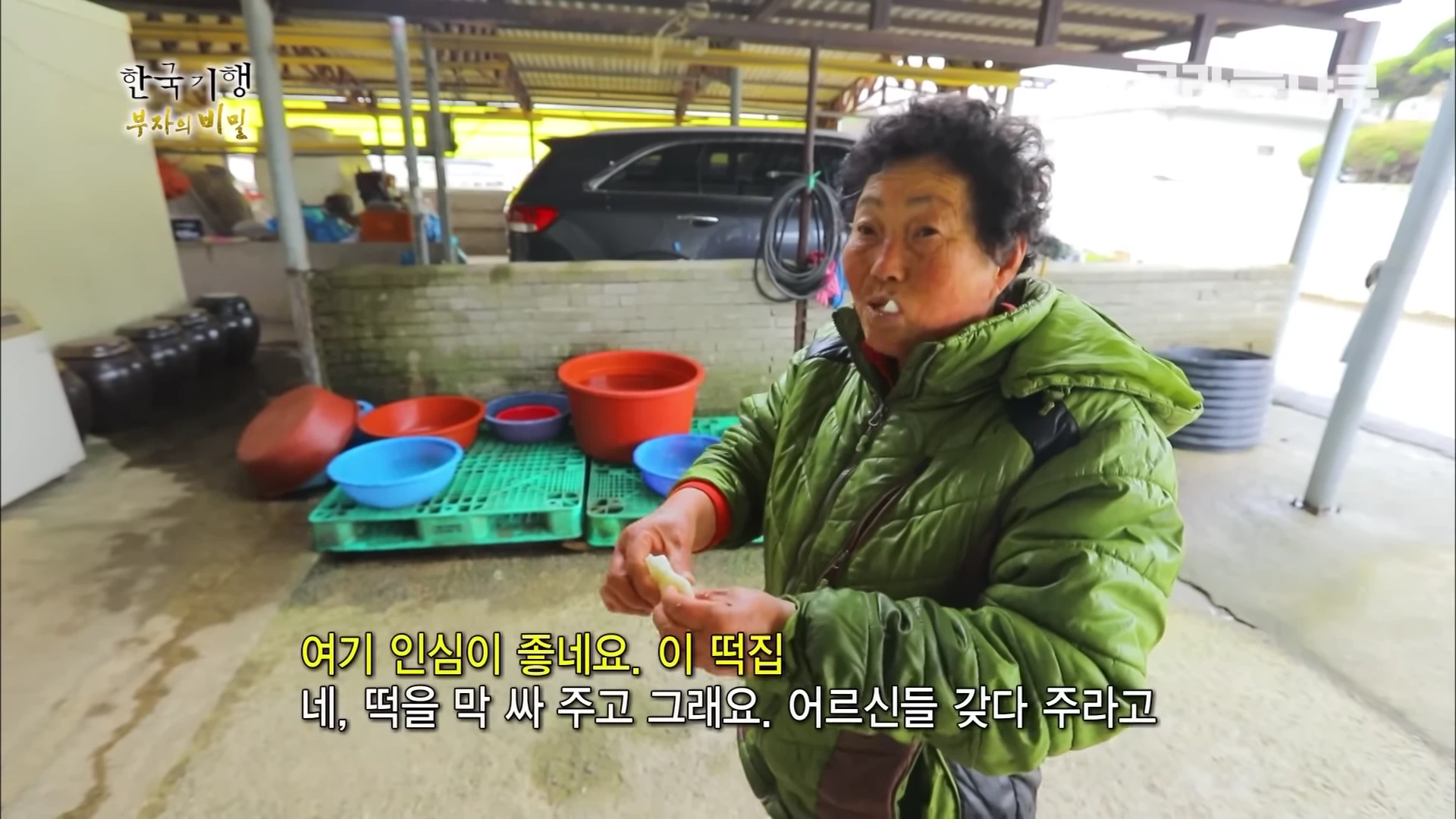 녹색 패딩을 입은 노년의 여성이 떡집 마당에서 떡을 먹으며 웃고 있는 모습