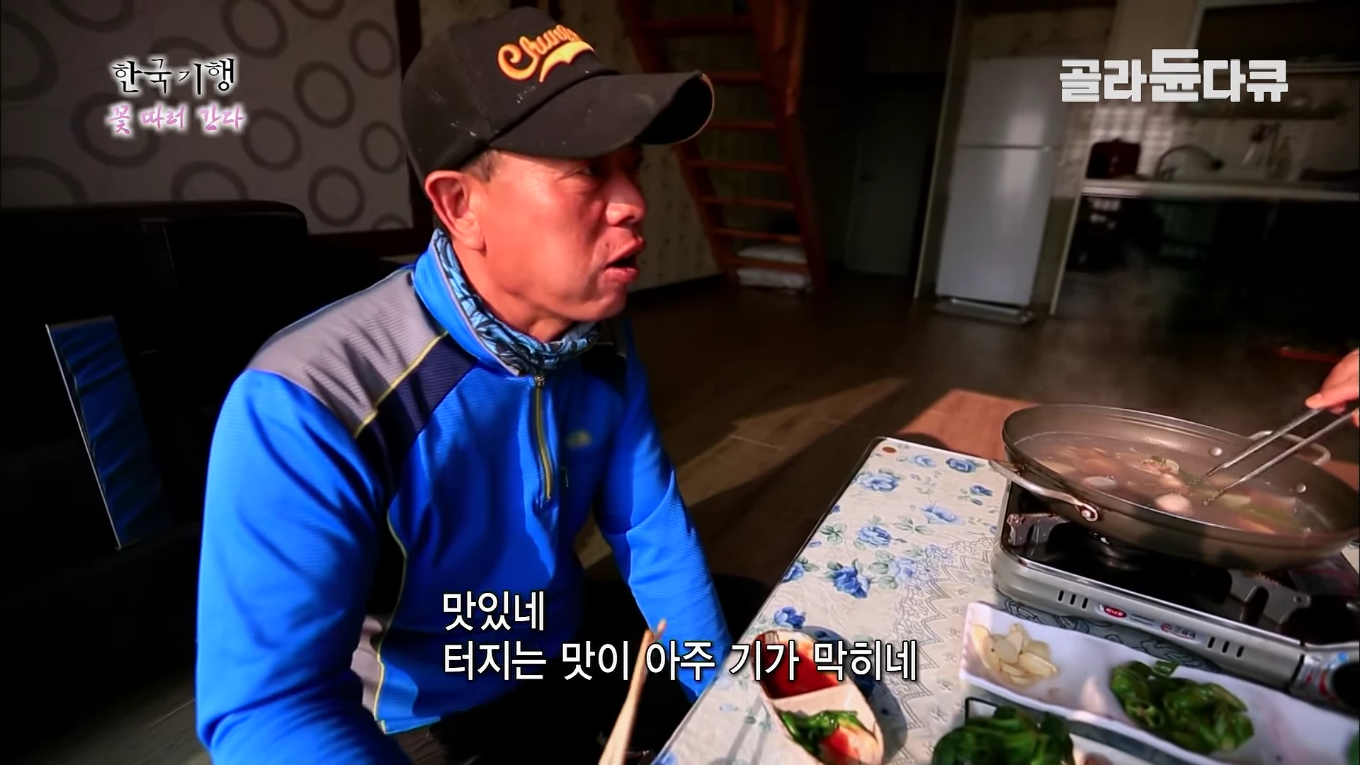 실내에서 검은색 야구 모자를 쓴 남성이 파란색 등산복을 입고 식탁에 앉아 주꾸미 요리를 먹으며 감탄하고 있다.