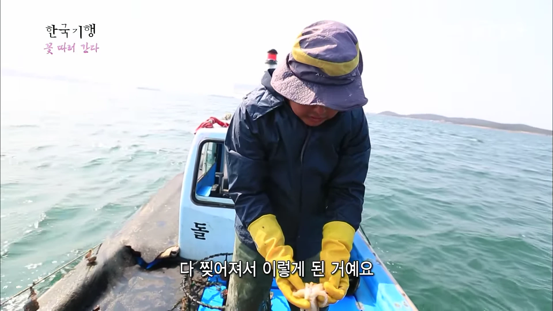 배 위에서 모자와 작업복을 입은 남성이 노란 고무장갑을 끼고 손에 잡힌 물체를 확인하고 있다. 화면 상단에는 방송 프로그램 로고와 자막이 표시되어 있다.