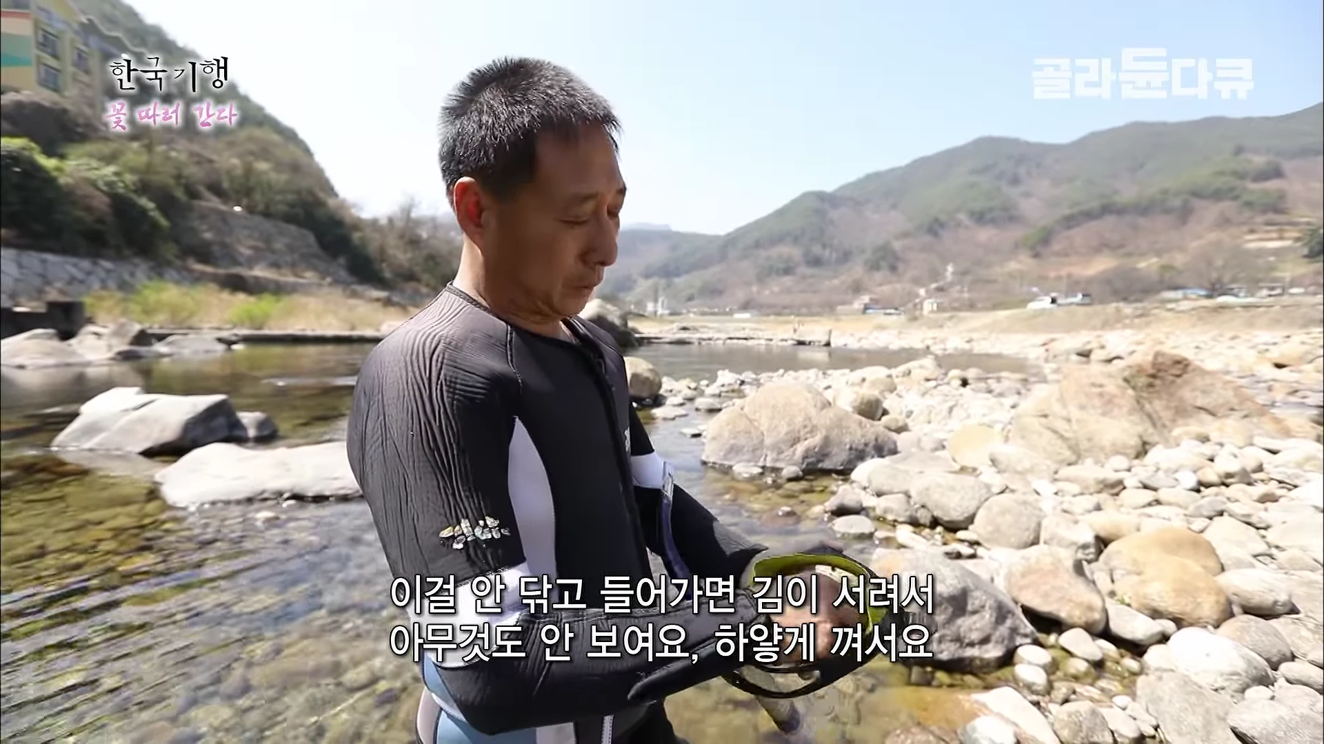 맑은 강가에서 잠수복을 입은 중년 남성이 물안경의 김 서림을 방지하기 위해 닦아내고 있는 모습이다.