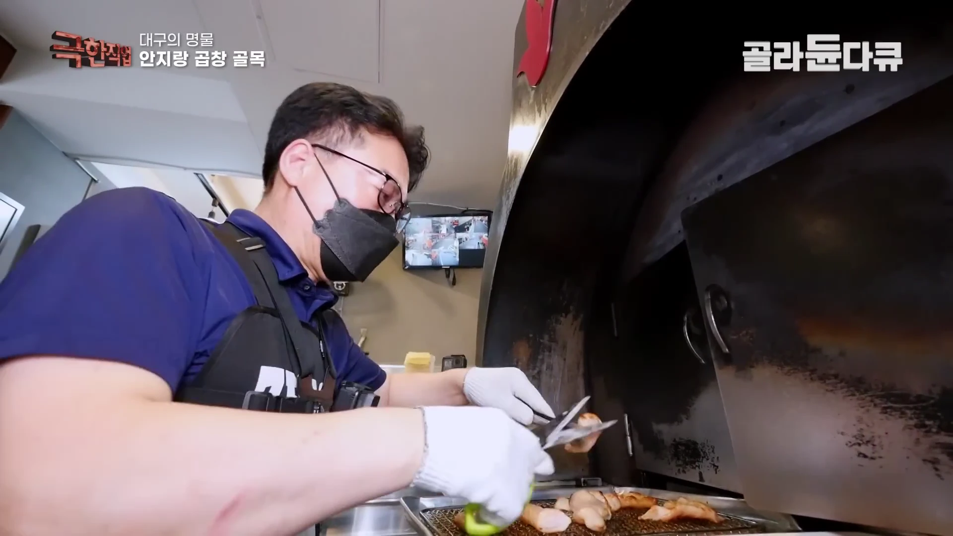 안경과 마스크를 쓴 남성이 앞치마를 두르고 화덕 앞에서 집게와 가위를 이용해 막창을 굽고 있는 모습