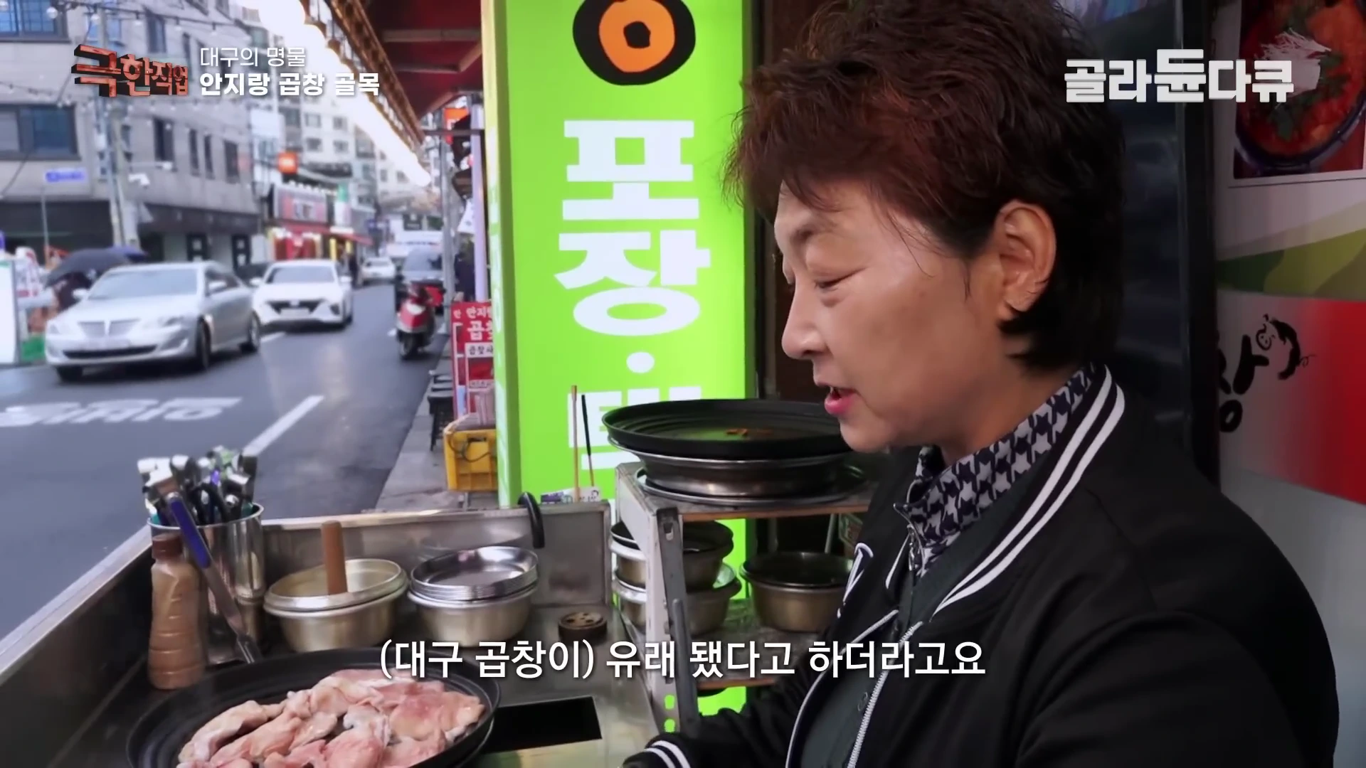 안지랑 곱창 골목의 한 식당 앞에서 중년 여성이 인터뷰하고 있으며, 그 옆에는 날 곱창이 담긴 철판이 놓여 있고 배경으로 식당가 거리가 보인다.