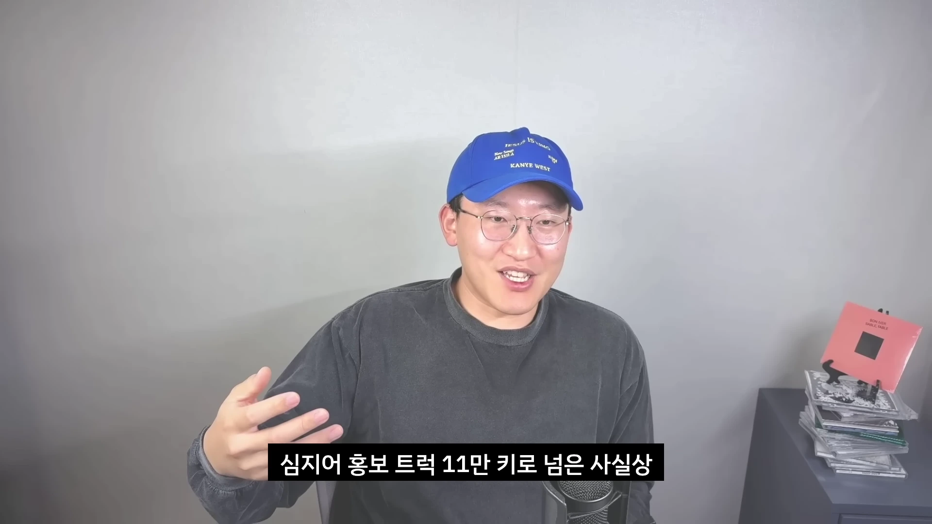 파란색 야구 모자를 쓴 남성이 마이크 앞에서 손동작을 하며 이야기를 하고 있는 모습.