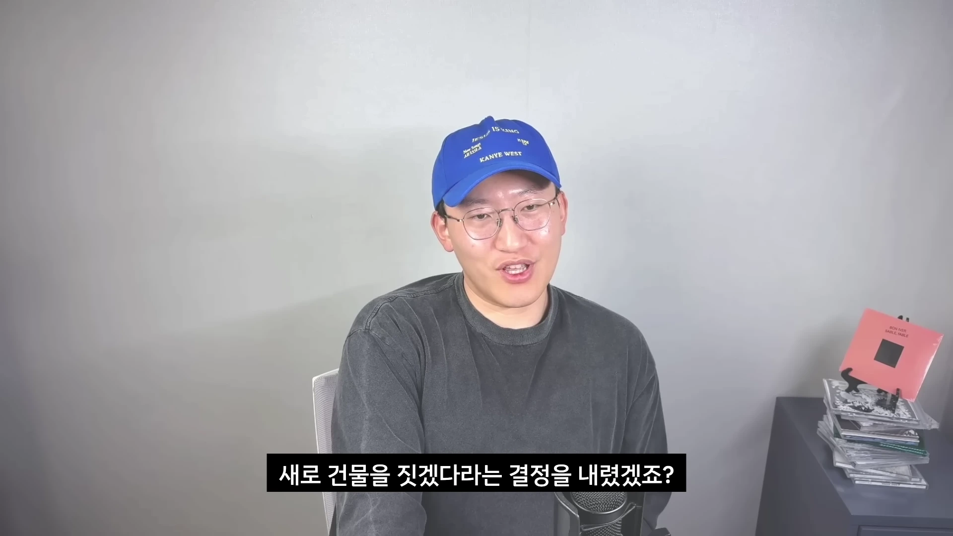 파란색 모자를 쓰고 안경을 쓴 남성이 밝은 회색 배경 앞에서 이야기를 하고 있으며, 하단에는 한국어 자막이 표시되어 있습니다.