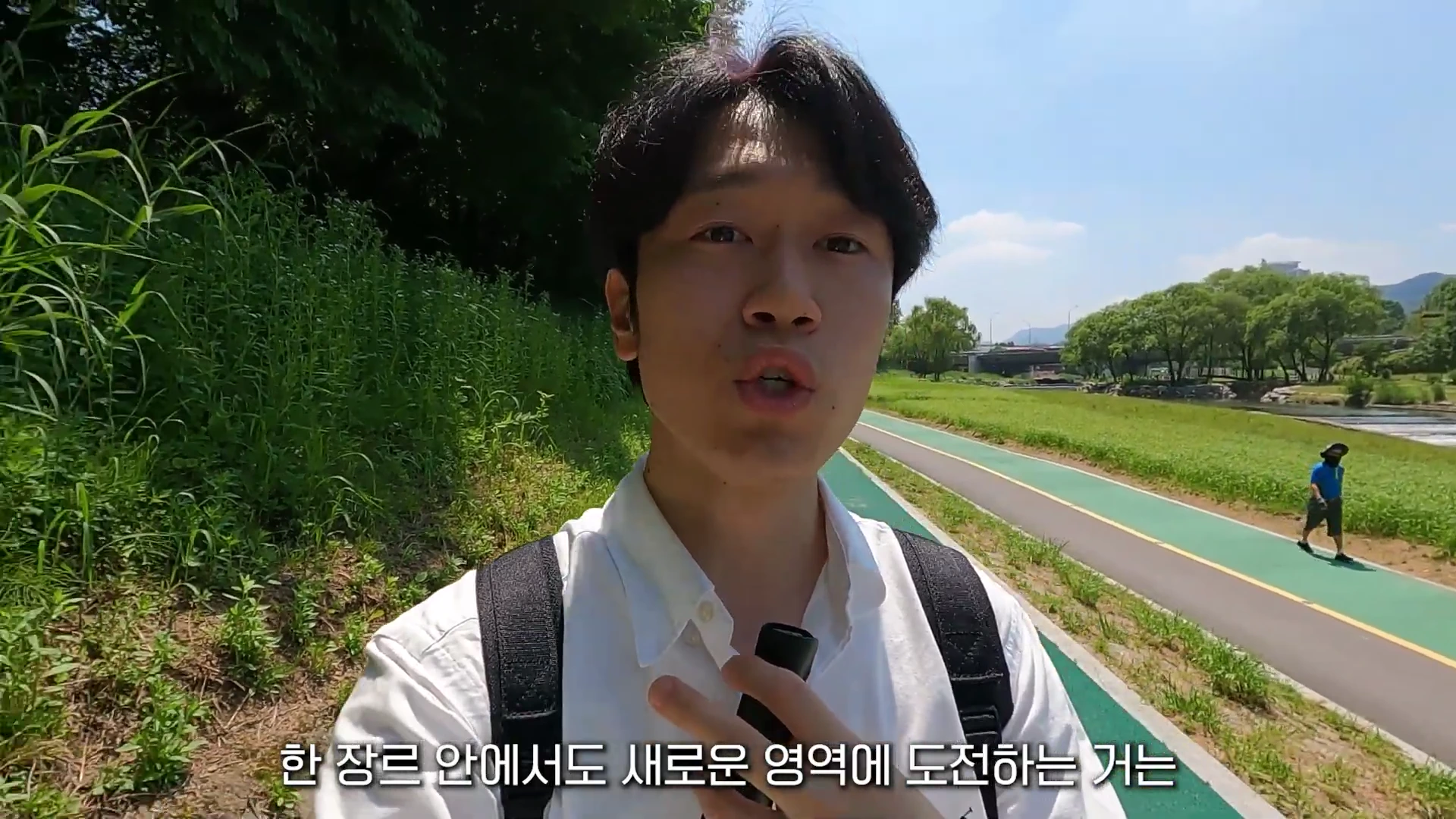 흰 셔츠를 입고 백팩을 멘 남성이 야외 산책로에서 카메라를 보며 이야기하고 있으며, 화면 하단에는 자막이 떠 있습니다.
