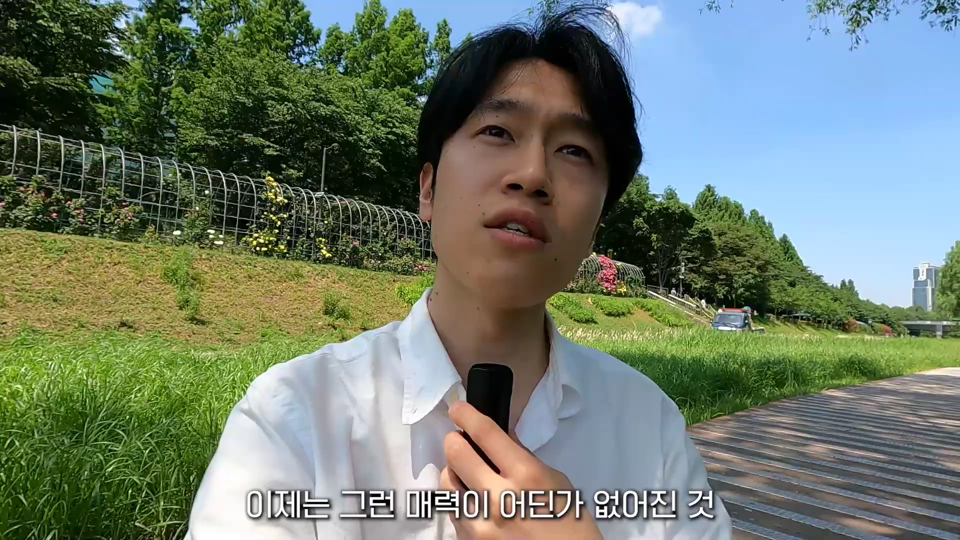 흰 셔츠를 입은 남성이 야외 공원에서 마이크를 들고 이야기하는 모습