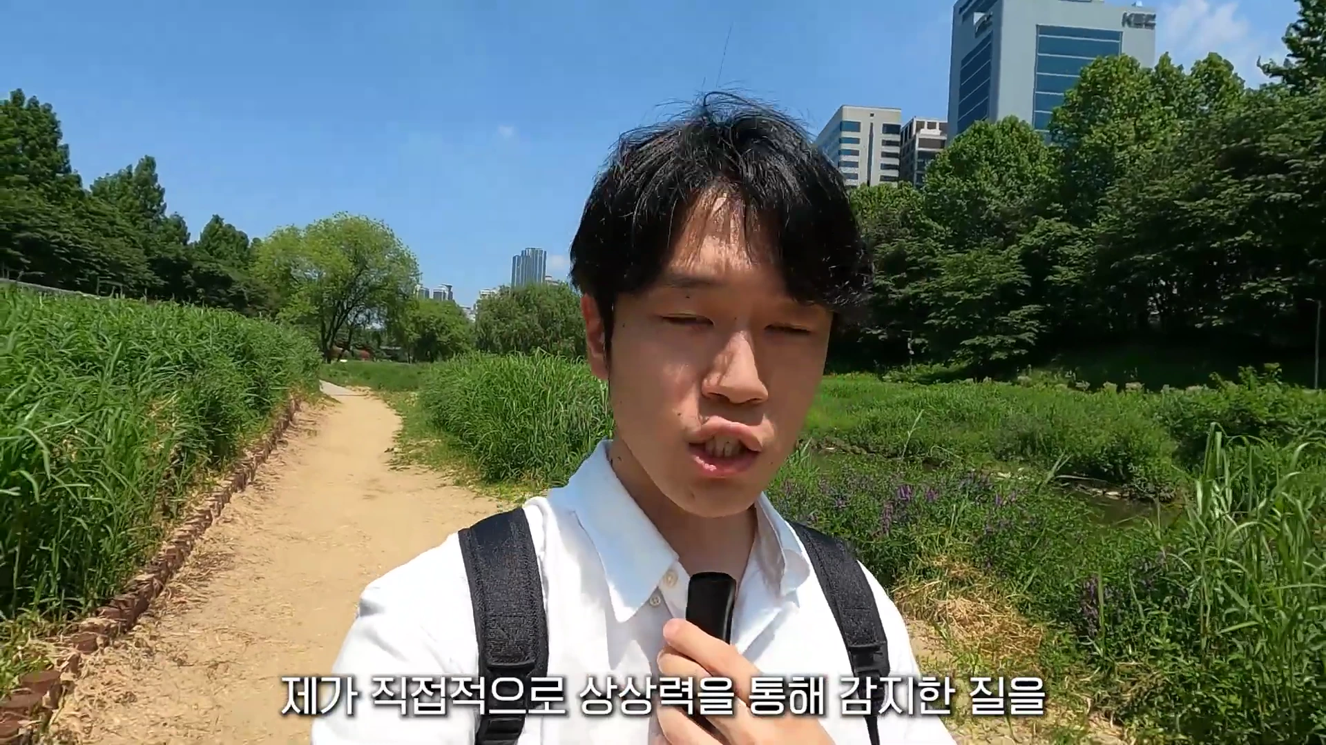 푸른 나무와 산책로가 보이는 양재천을 배경으로 흰 셔츠를 입은 남성이 카메라를 보며 이야기하고 있는 모습