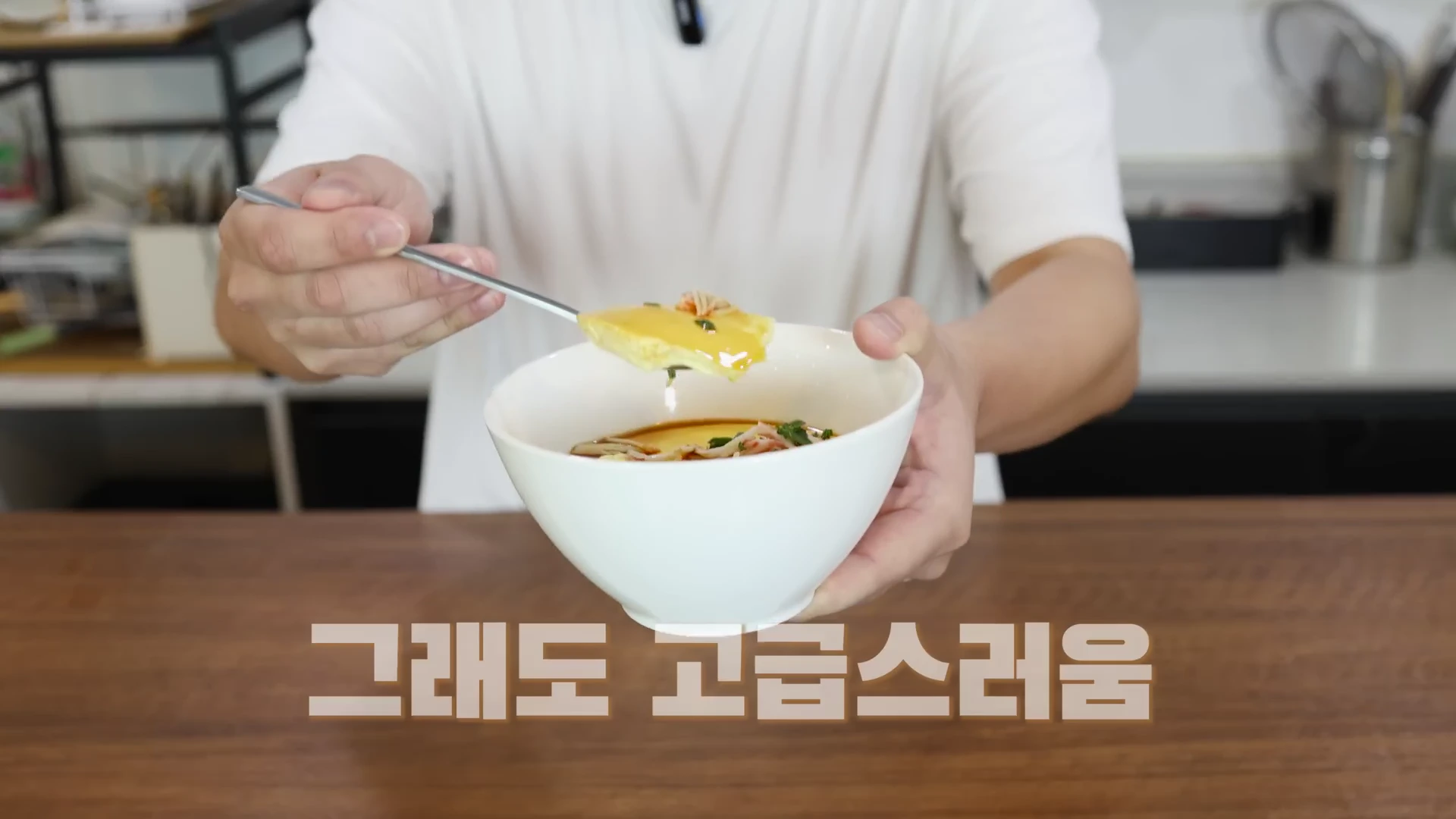 흰색 티셔츠를 입은 사람이 푸딩처럼 부드러운 계란찜을 숟가락으로 떠서 보여주고 있으며, 화면 하단에는 '그래도 고급스러움'이라는 한글 자막이 적혀 있습니다.