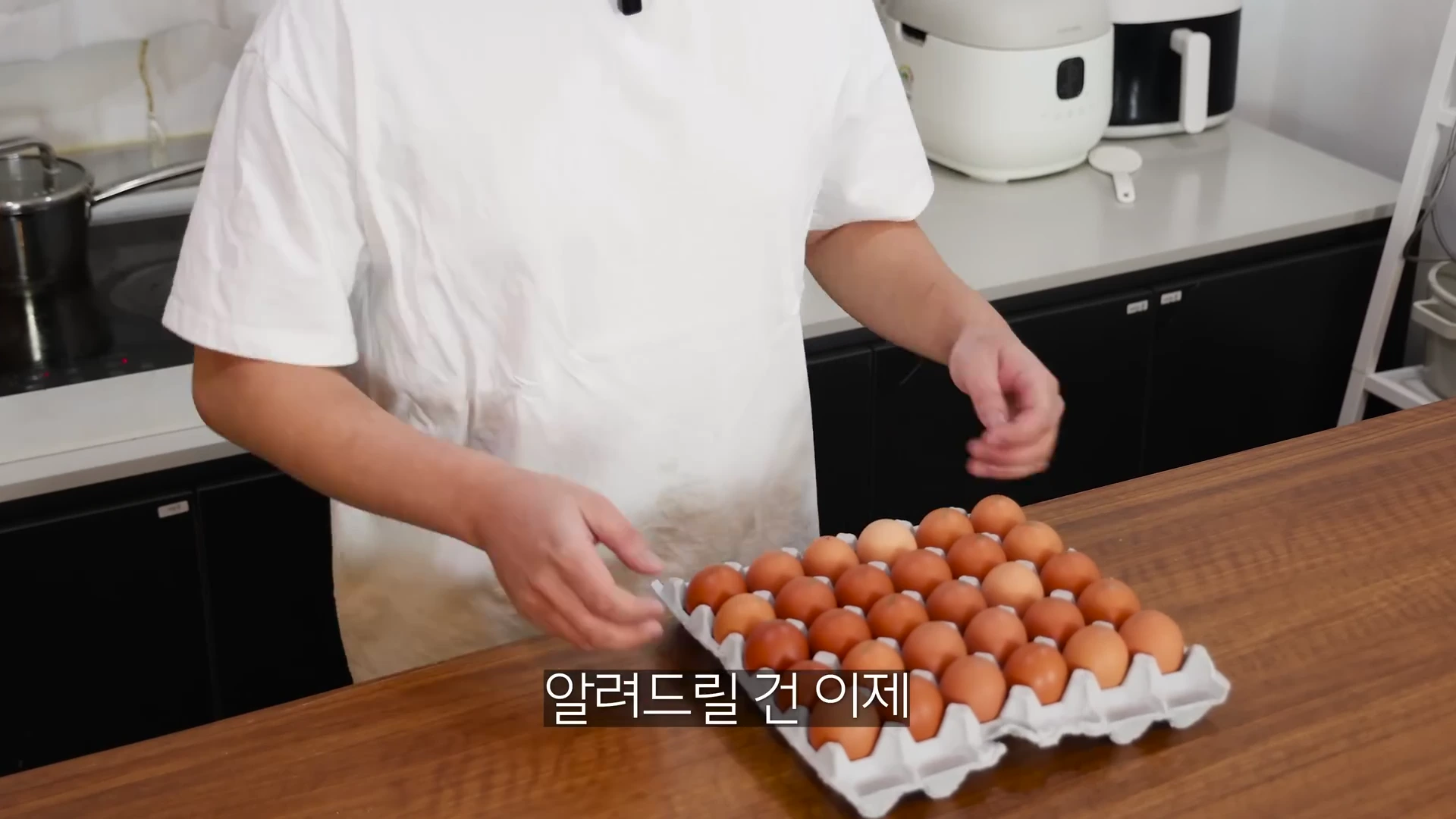 흰 티셔츠를 입은 사람이 주방 식탁 위에 놓인 계란 한 판 앞에서 요리에 대해 설명하고 있는 모습
