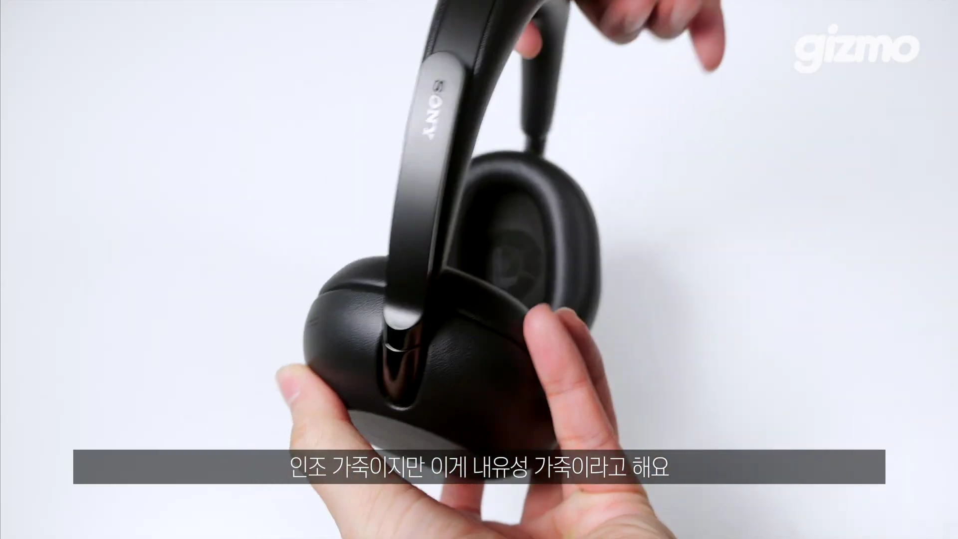 손으로 검은색 무선 헤드폰의 이어컵과 프레임을 잡고 보여주고 있으며 화면 하단에는 인조 가죽 소재에 대한 한국어 자막이 표시되어 있습니다.