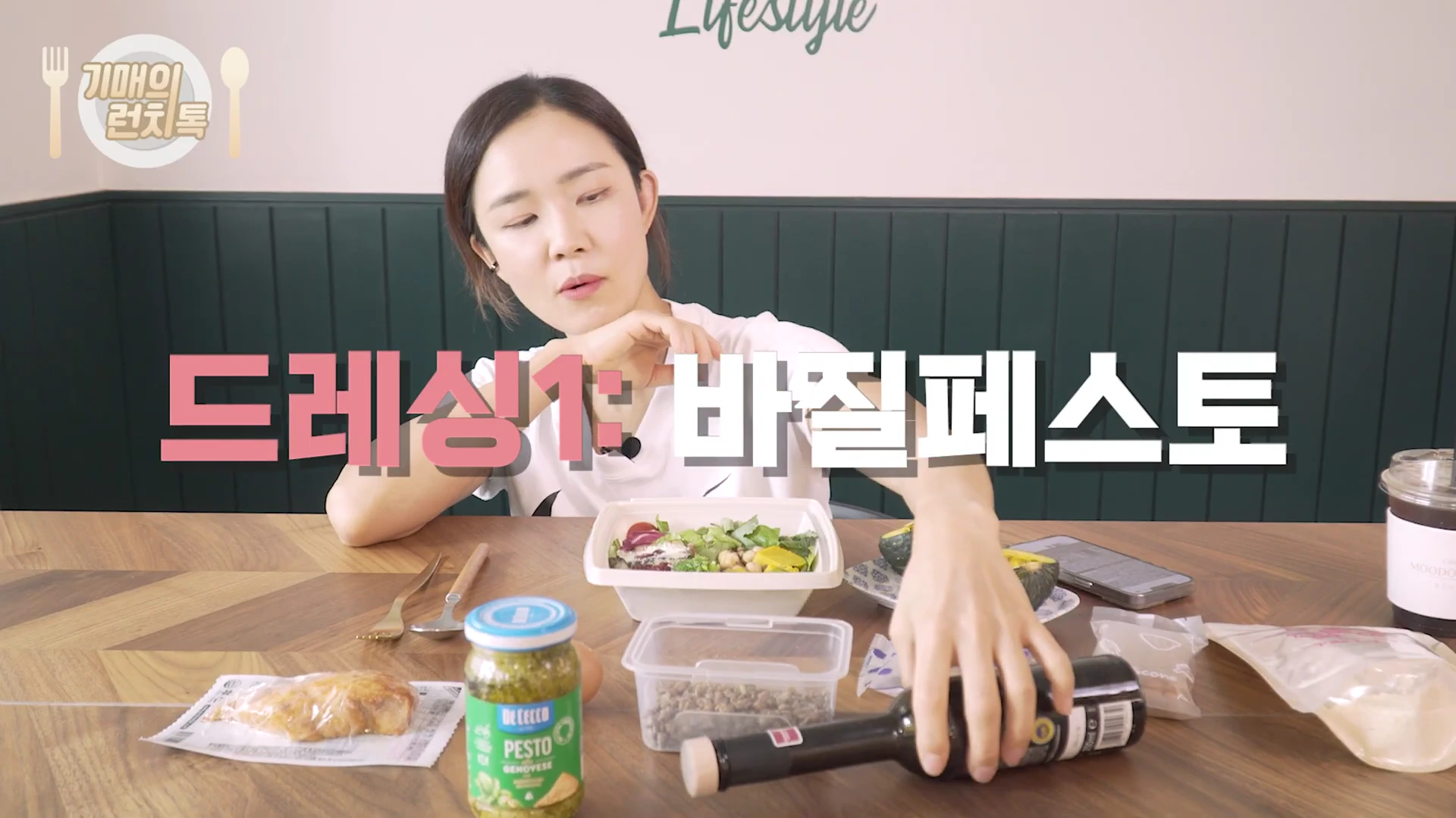 흰색 티셔츠를 입은 여성이 테이블 앞에 앉아 샐러드와 바질 페스토 병, 발사믹 식초 병을 앞에 두고 이야기하고 있는 모습