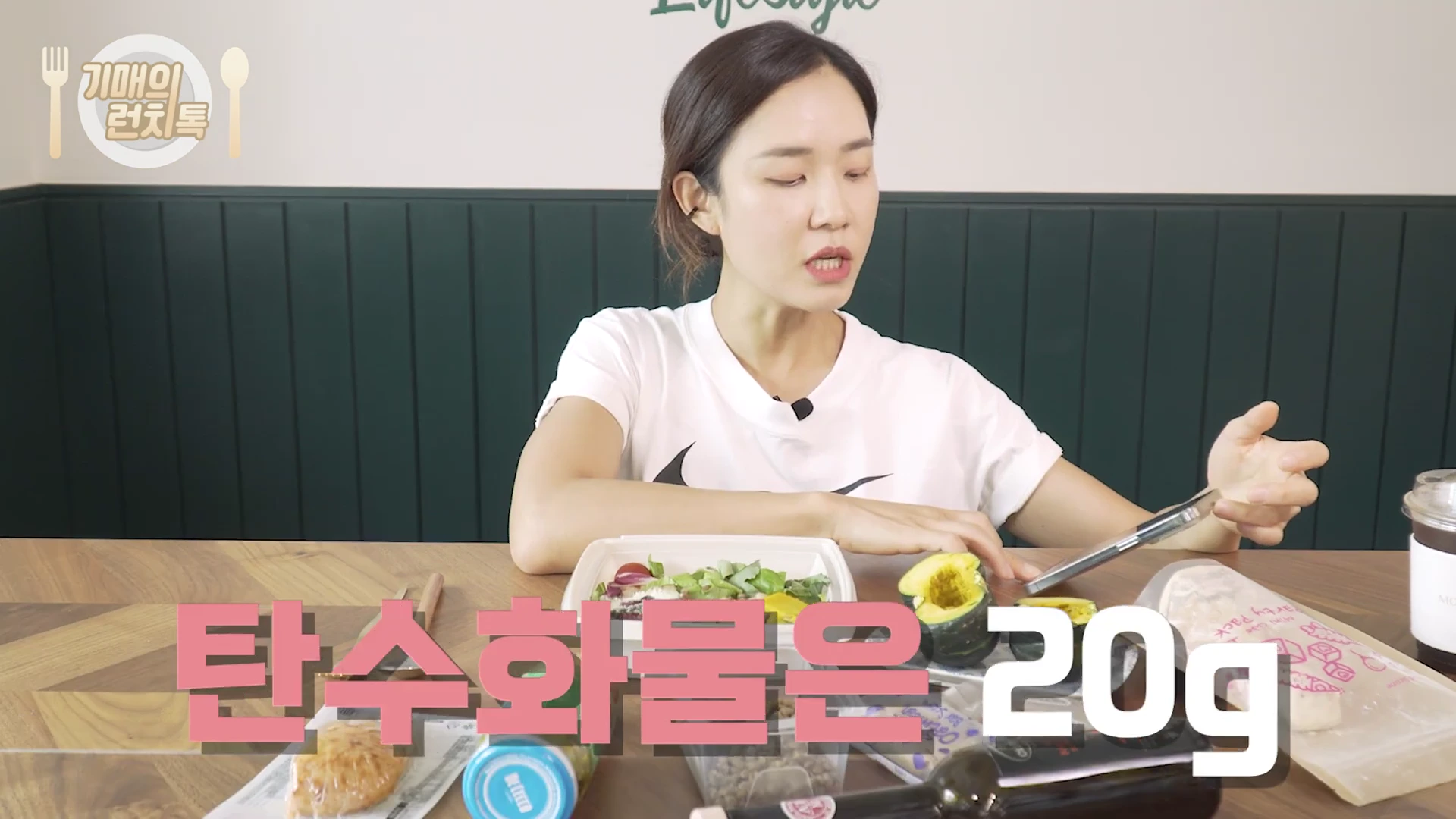 흰색 반팔 티셔츠를 입은 여성이 테이블에 앉아 스마트폰을 보며 대화하고 있으며, 화면 하단에는 핑크색 자막으로 '탄수화물은 20g'이라는 글자가 크게 적혀 있습니다.