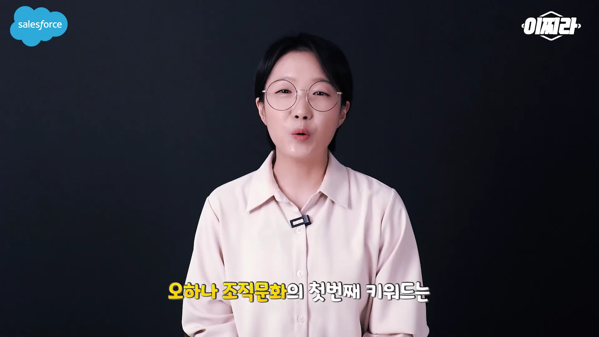안경을 쓴 여성 진행자가 검은색 배경 앞에서 오하나 조직문화에 대해 설명하고 있는 영상의 한 장면입니다.