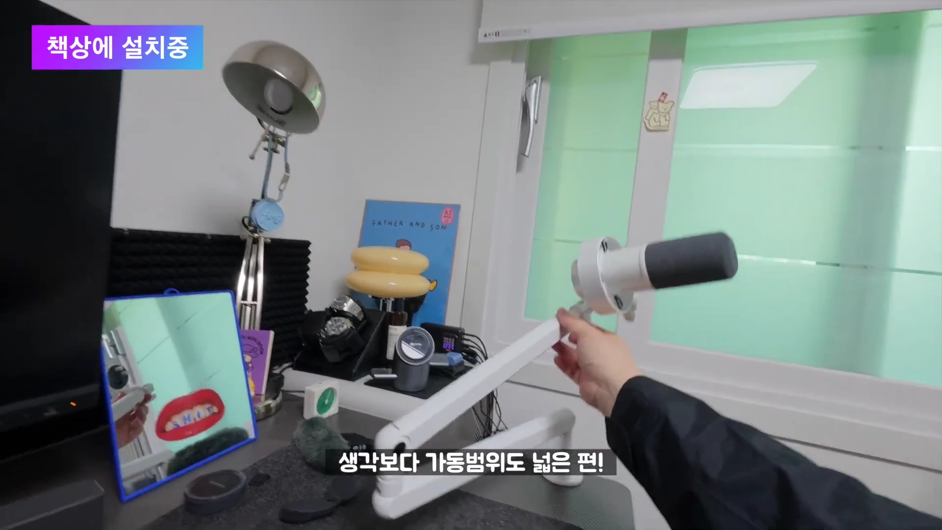 실내 책상 위에서 한 손으로 마이크가 장착된 흰색 붐암 스탠드를 움직이고 있는 모습이며 화면 왼쪽 상단에는 '책상에 설치중'이라는 자막이 있다.