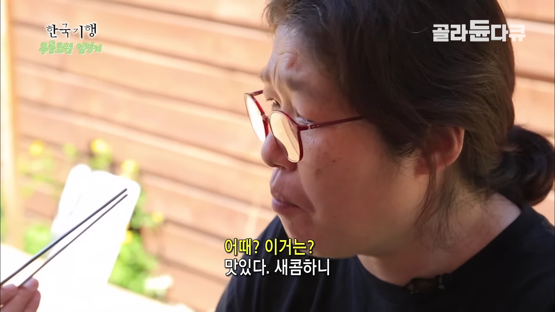 붉은색 테 안경을 쓴 중년 여성이 젓가락으로 음식을 먹으며 맛을 음미하고 있는 모습.