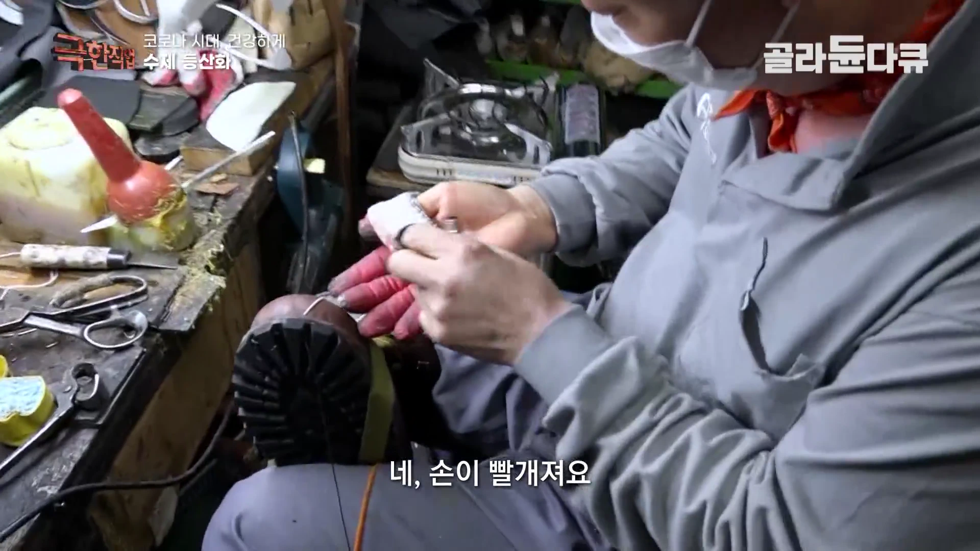 작업대에 앉은 장인이 붉은 장갑을 낀 손으로 등산화 밑창에 실을 꿰고 있는 모습이며, 주변에는 각종 도구와 재료들이 놓여 있습니다.
