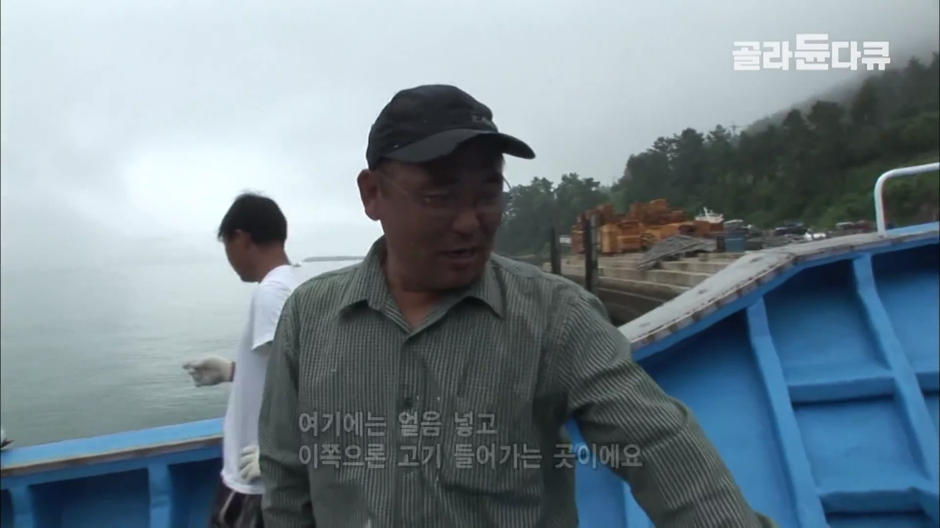 배 위에서 검은색 모자를 쓴 남성이 환하게 웃으며 자신이 서 있는 공간의 용도를 설명하고 있고, 그 뒤로 또 다른 작업자가 서 있다.