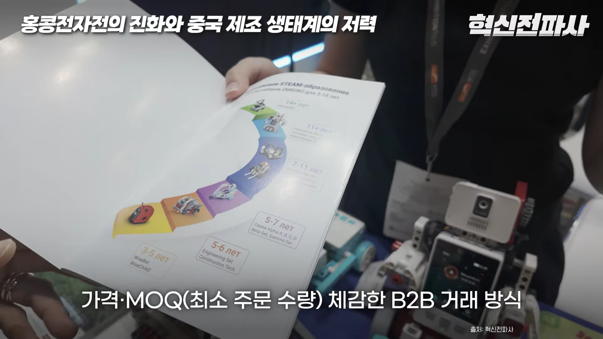사람의 손이 들고 있는 로봇 교육 제품 카탈로그 페이지에 연령별 제품 구성표가 인쇄되어 있으며, 배경에는 로봇 부품이 일부 보입니다.