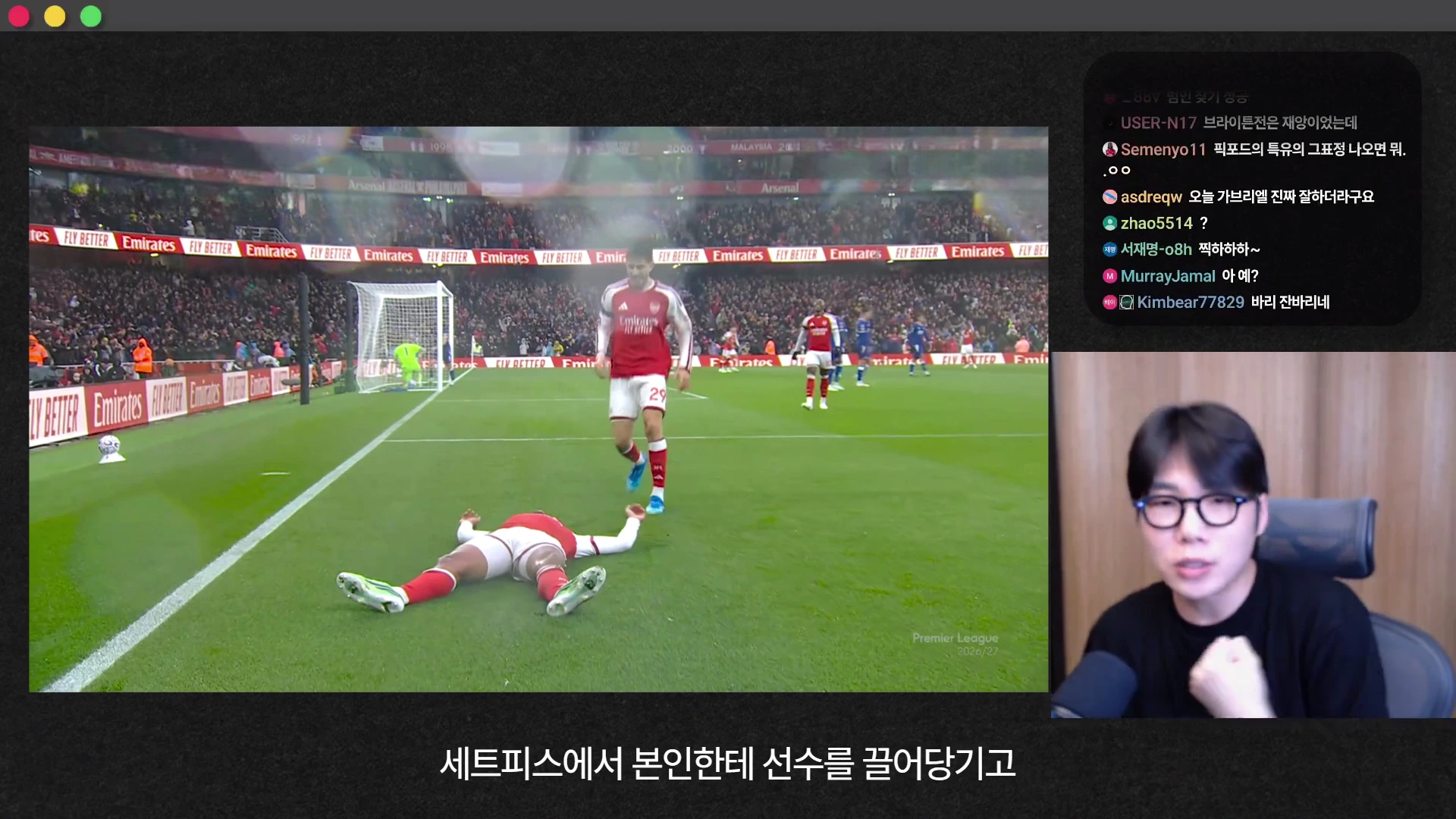 축구 경기장에서 한 선수가 그라운드에 누워있고 다른 선수가 그 옆을 지나가는 모습이 담긴 경기 영상 화면과, 그 옆으로 방송을 진행하는 안경 쓴 남성의 모습이 담긴 화면