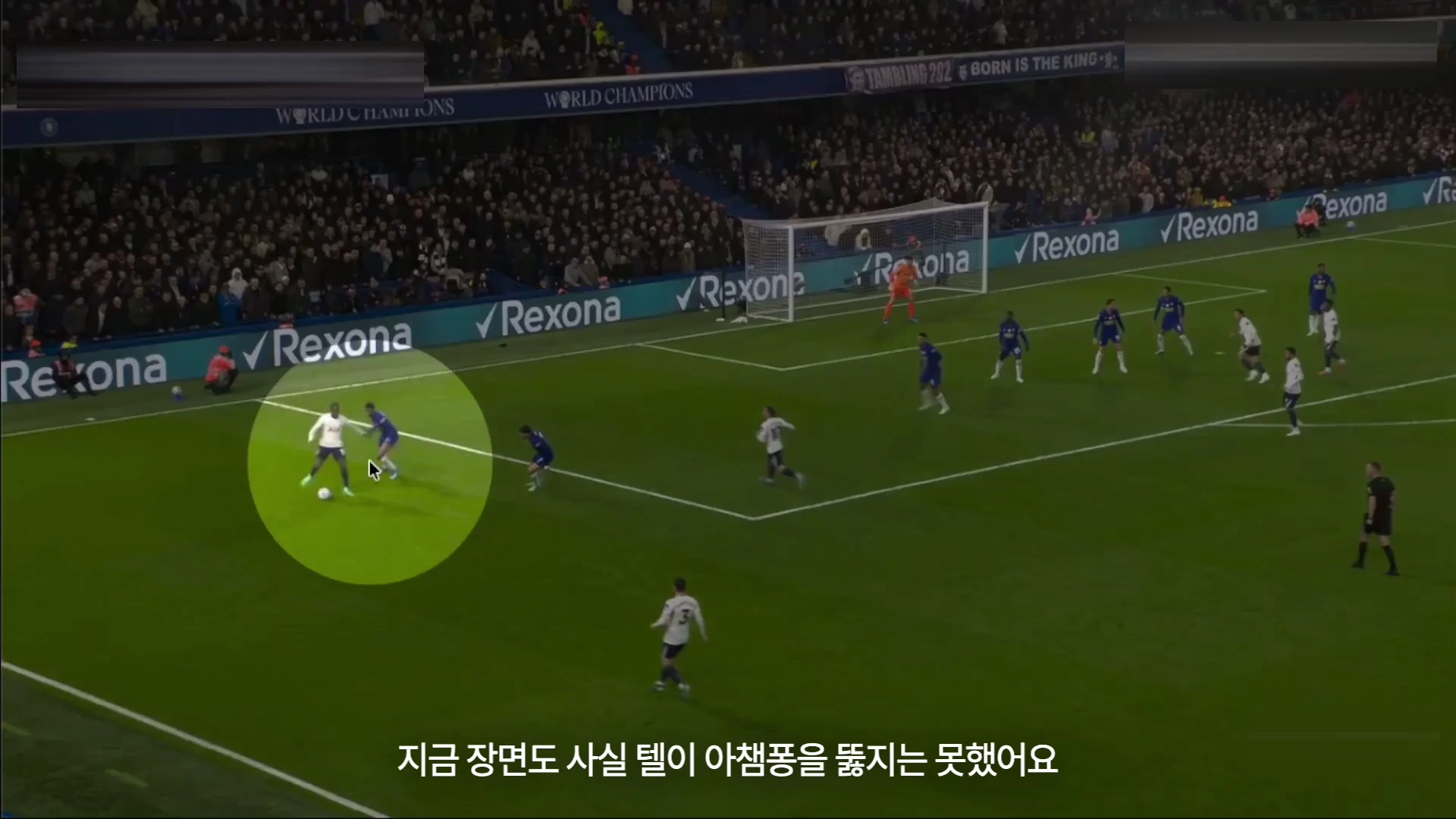 축구 경기장에서 흰색 유니폼을 입은 공격수가 노란색 원으로 강조된 상태에서 상대 수비수와 대치하며 공을 몰고 가고 있습니다.