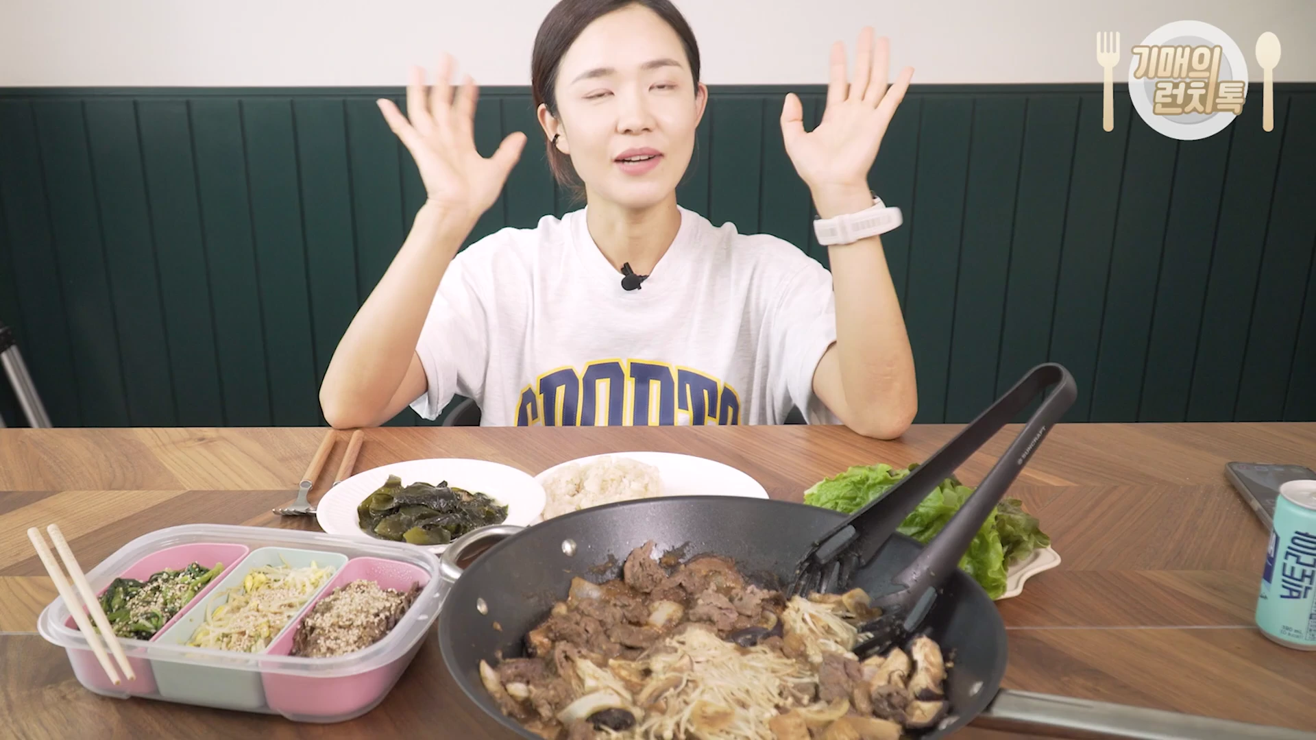 밝은 실내에서 한 여성이 책상 앞에 앉아 양손을 들어 올리며 이야기를 나누고 있으며, 앞에는 불고기와 나물 반찬, 밥이 놓여 있습니다.