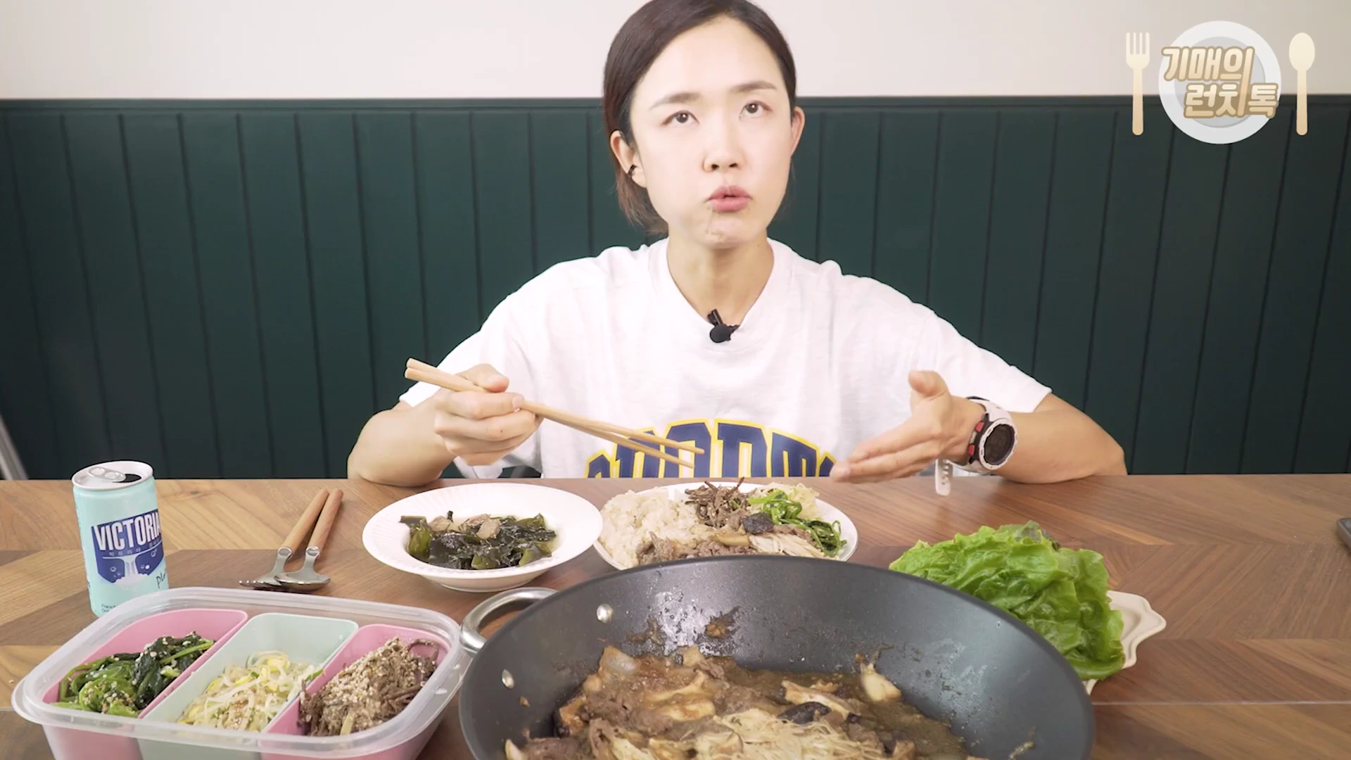 흰색 티셔츠를 입은 여성이 식탁에 앉아 나물과 불고기 등 한식 반찬을 앞에 두고 이야기를 나누고 있는 모습