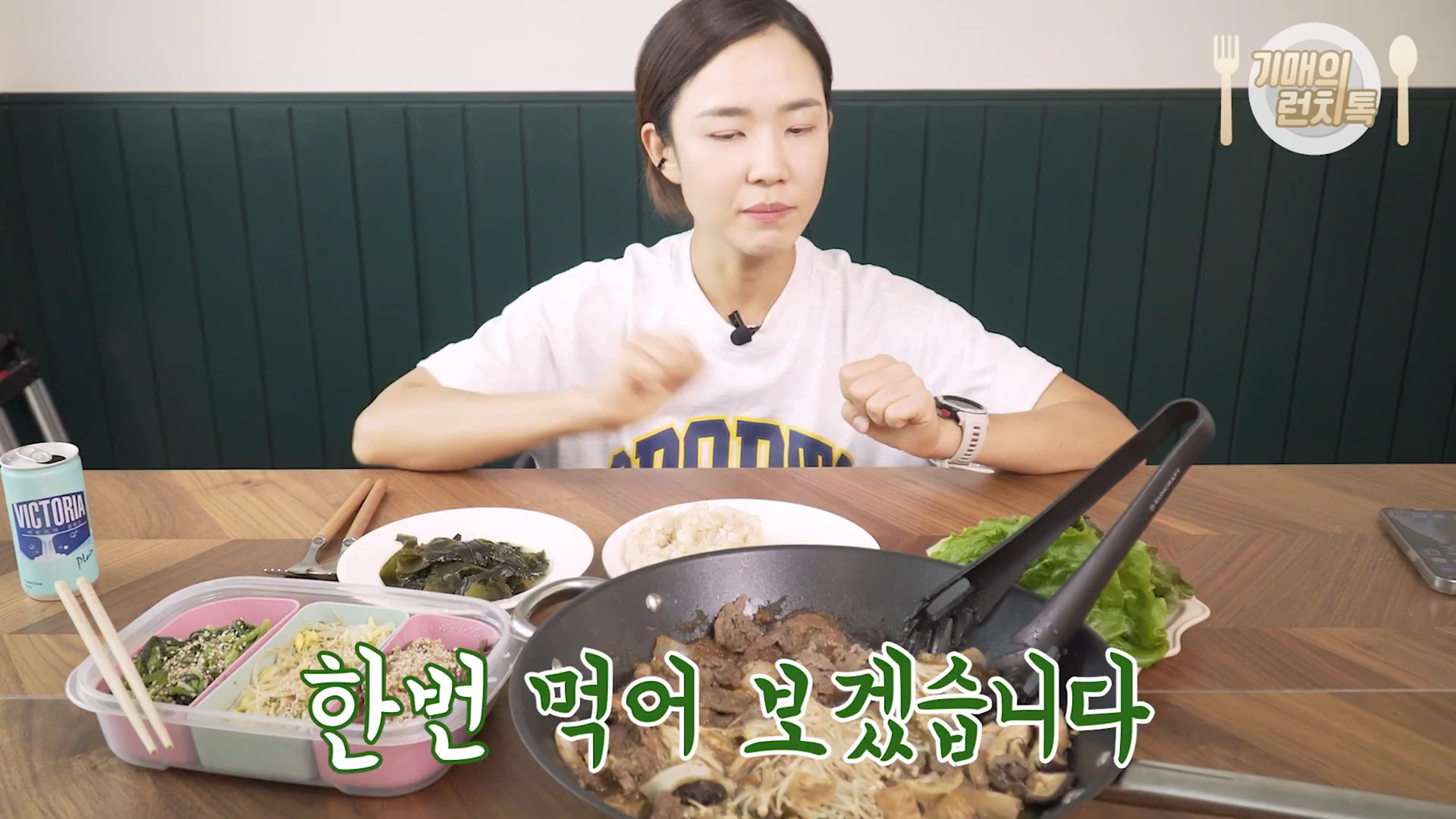 흰색 티셔츠를 입은 여성이 식탁 앞에 앉아 있고, 앞에는 불고기 팬, 나물 반찬통, 밥그릇, 물이 놓여 있는 영상 화면.