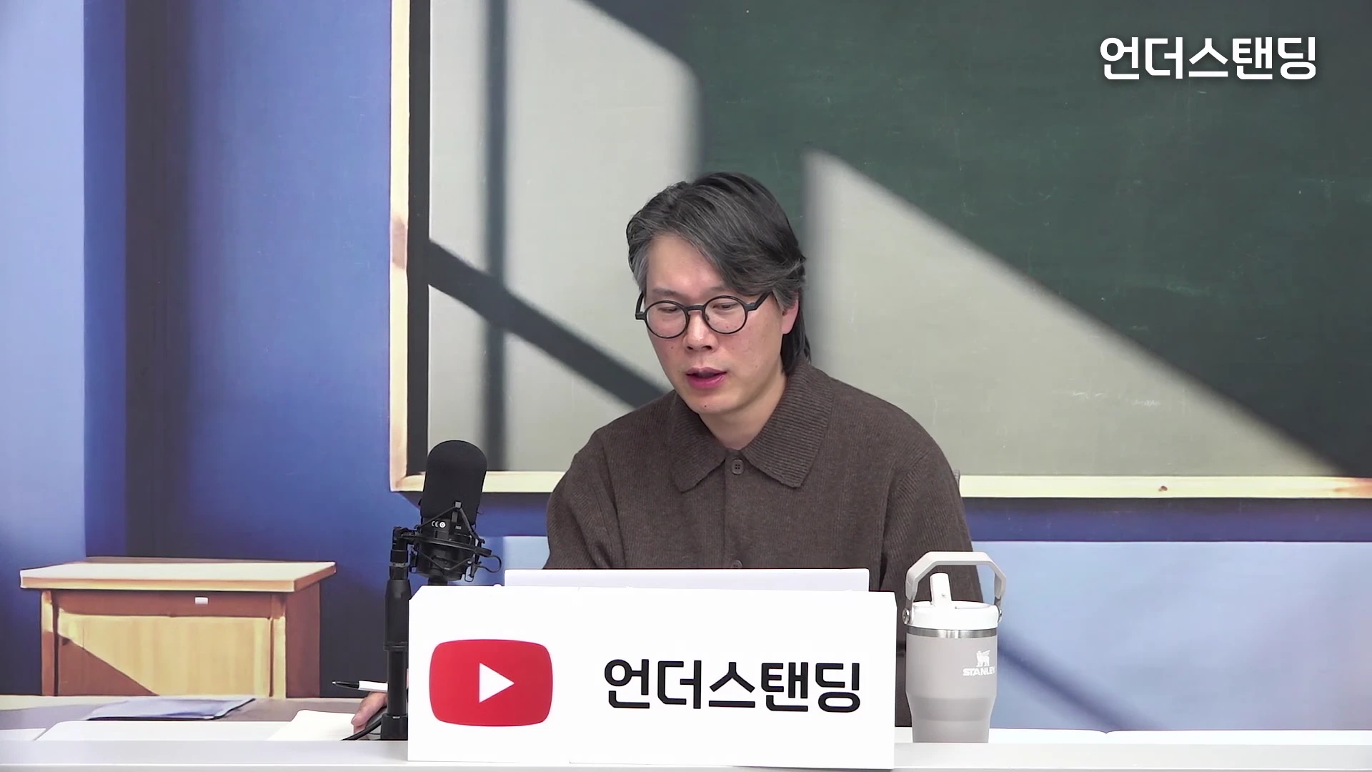 안경을 쓴 남성이 스튜디오 테이블 앞에 앉아 마이크를 앞에 두고 이야기를 나누고 있다.