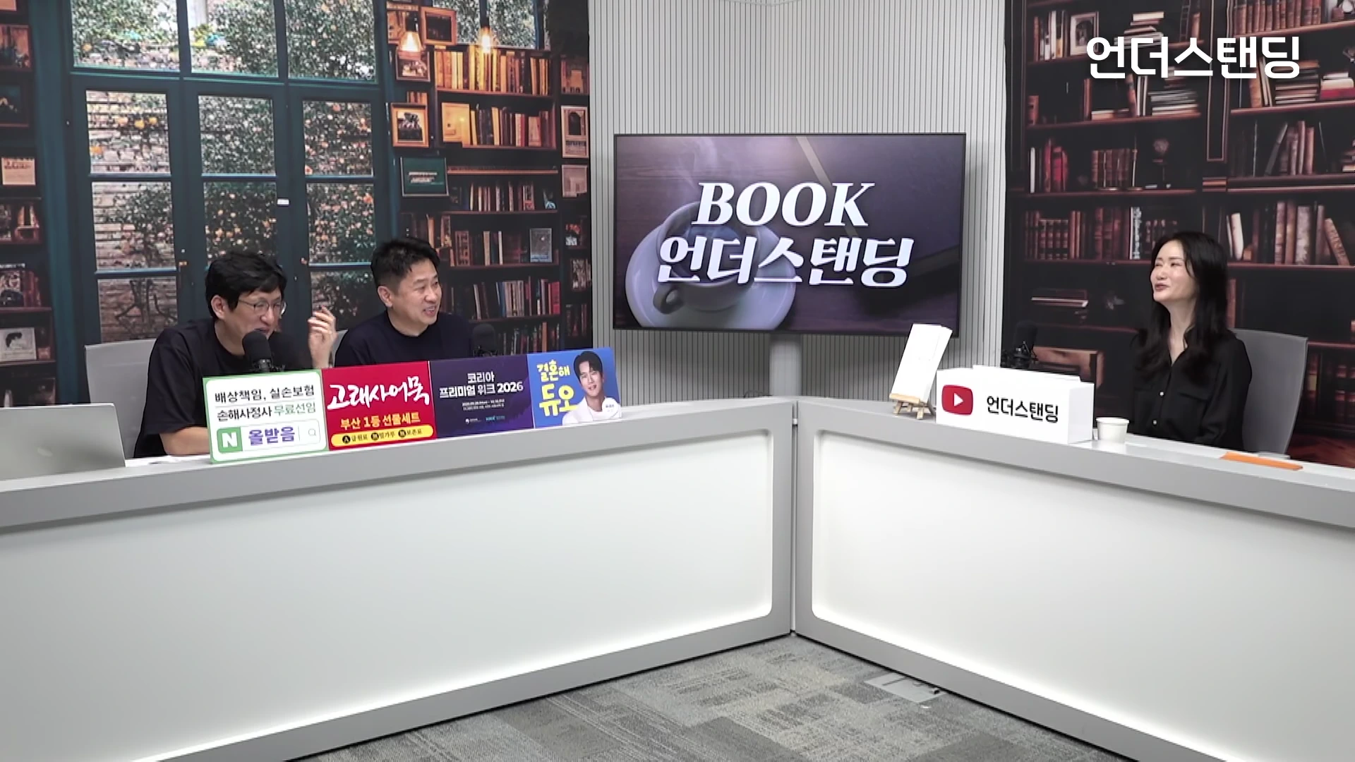 두 명의 남성과 한 명의 여성이 스튜디오 테이블에 앉아 대화를 나누고 있는 모습. 배경에는 책장이 그려진 벽면과 'BOOK 언더스탠딩' 로고가 표시된 대형 화면이 있다.