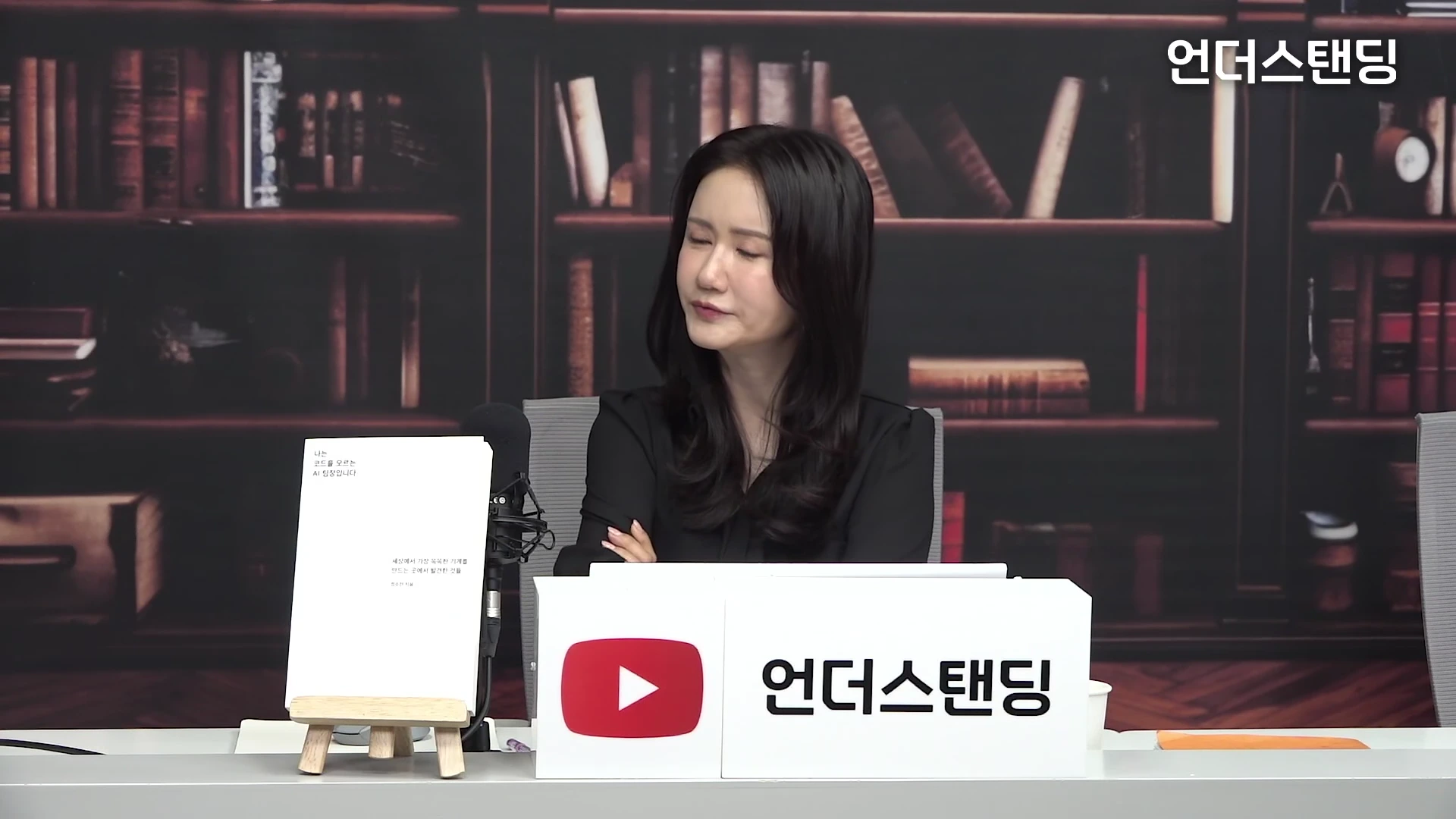 서재 배경의 스튜디오에서 검은 옷을 입은 여성이 팔짱을 끼고 앉아 있고, 그 앞에는 책 한 권이 거치대에 놓여 있다.