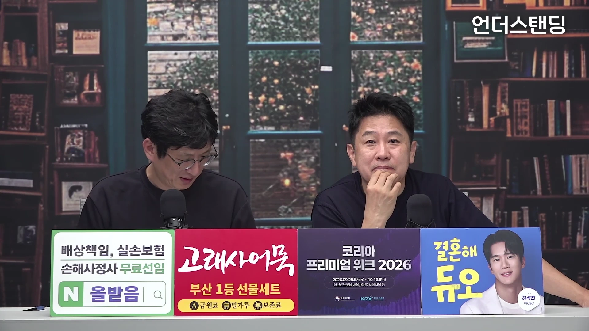 두 남자가 스튜디오에서 마이크 앞에 앉아 대화를 나누고 있으며, 책장 배경의 벽면 앞에 테이블이 놓여 있고 테이블 전면에는 네 가지 색상의 홍보 배너가 부착되어 있습니다.