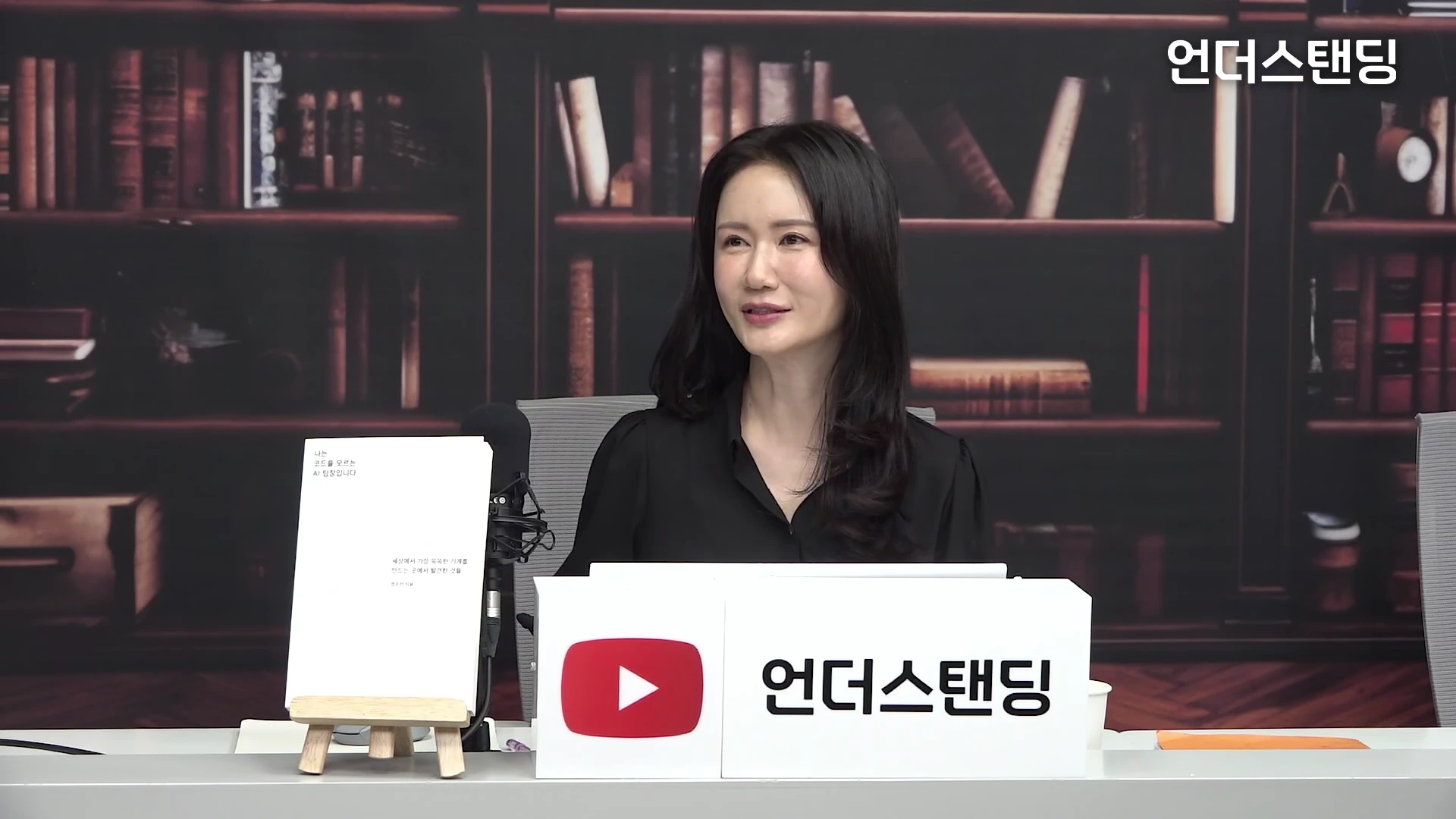 검은색 셔츠를 입은 여성 출연자가 책이 올려진 책상 앞에 앉아 이야기하고 있는 모습. 배경에는 책장이 있으며, 책상 앞에는 유튜브 로고와 언더스탠딩 로고가 새겨진 흰색 표지판이 놓여 있다.