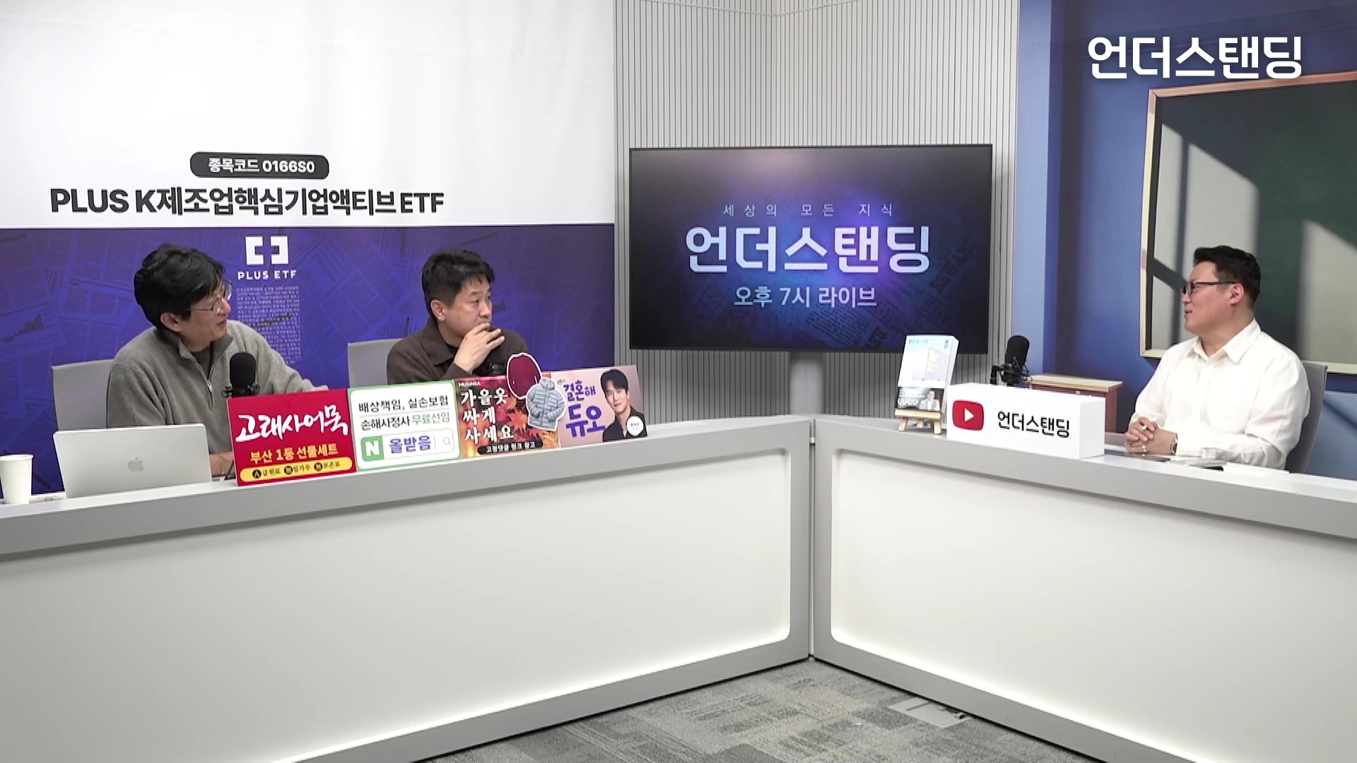 밝은 스튜디오 세트에서 세 명의 남성이 마주 앉아 대화를 나누는 모습입니다. 앞에는 흰색 책상과 마이크, 홍보 문구가 적힌 패널들이 놓여 있습니다.
