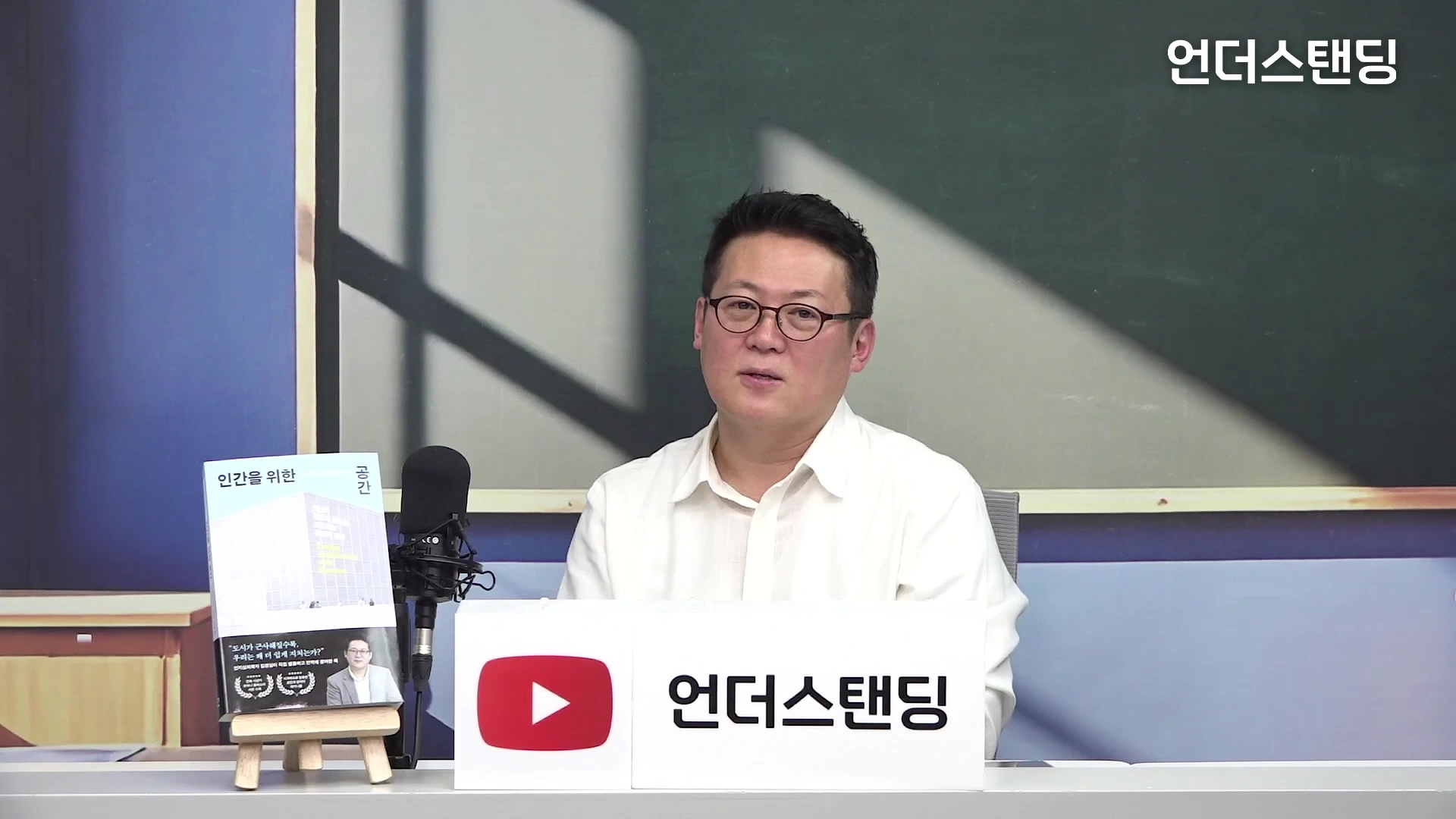 흰색 셔츠를 입고 안경을 쓴 남성이 스튜디오 책상에 앉아 이야기하고 있으며, 책상 위에는 마이크와 책이 놓여 있고 배경에는 칠판 같은 보드와 유튜브 로고가 보입니다.