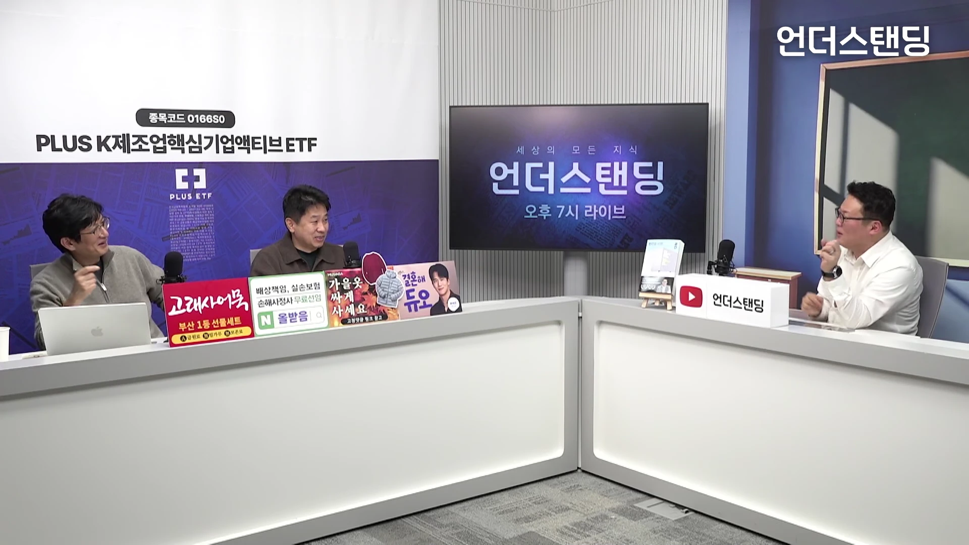 두 명의 진행자가 마주 보고 앉아 대화를 나누는 스튜디오 전경. 화이트 톤의 데스크와 대형 디스플레이 화면이 배치되어 있고, 테이블 위에는 다양한 상품 홍보 문구가 적힌 피켓들이 놓여 있다.