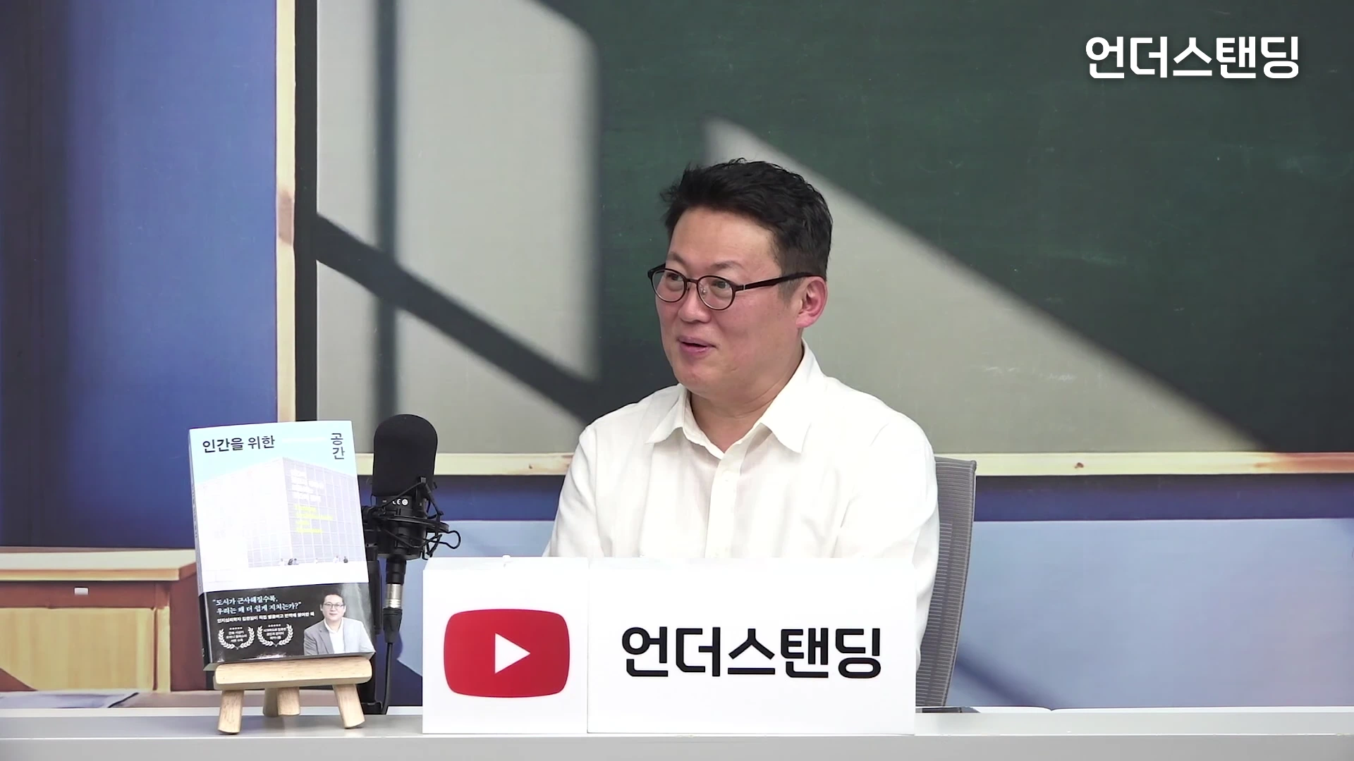 안경을 쓴 남성이 스튜디오에서 마이크 앞에 앉아 웃으며 대화하고 있으며, 옆에는 '인간을 위한 공간'이라는 책이 놓여 있습니다.