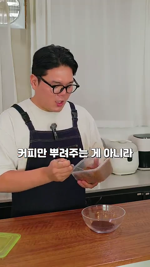 안경을 쓴 남성이 앞치마를 두르고 주방에서 투명한 용기에 담긴 갈색 가루를 숟가락으로 젓고 있는 모습.