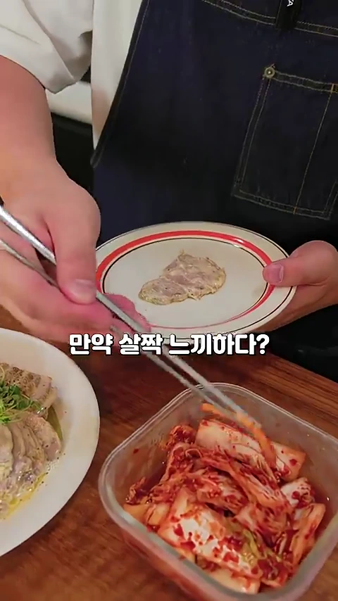 앞치마를 입은 요리사가 젓가락으로 김치를 집어 우유에 졸인 고기 요리 접시 위에 올리고 있는 모습.