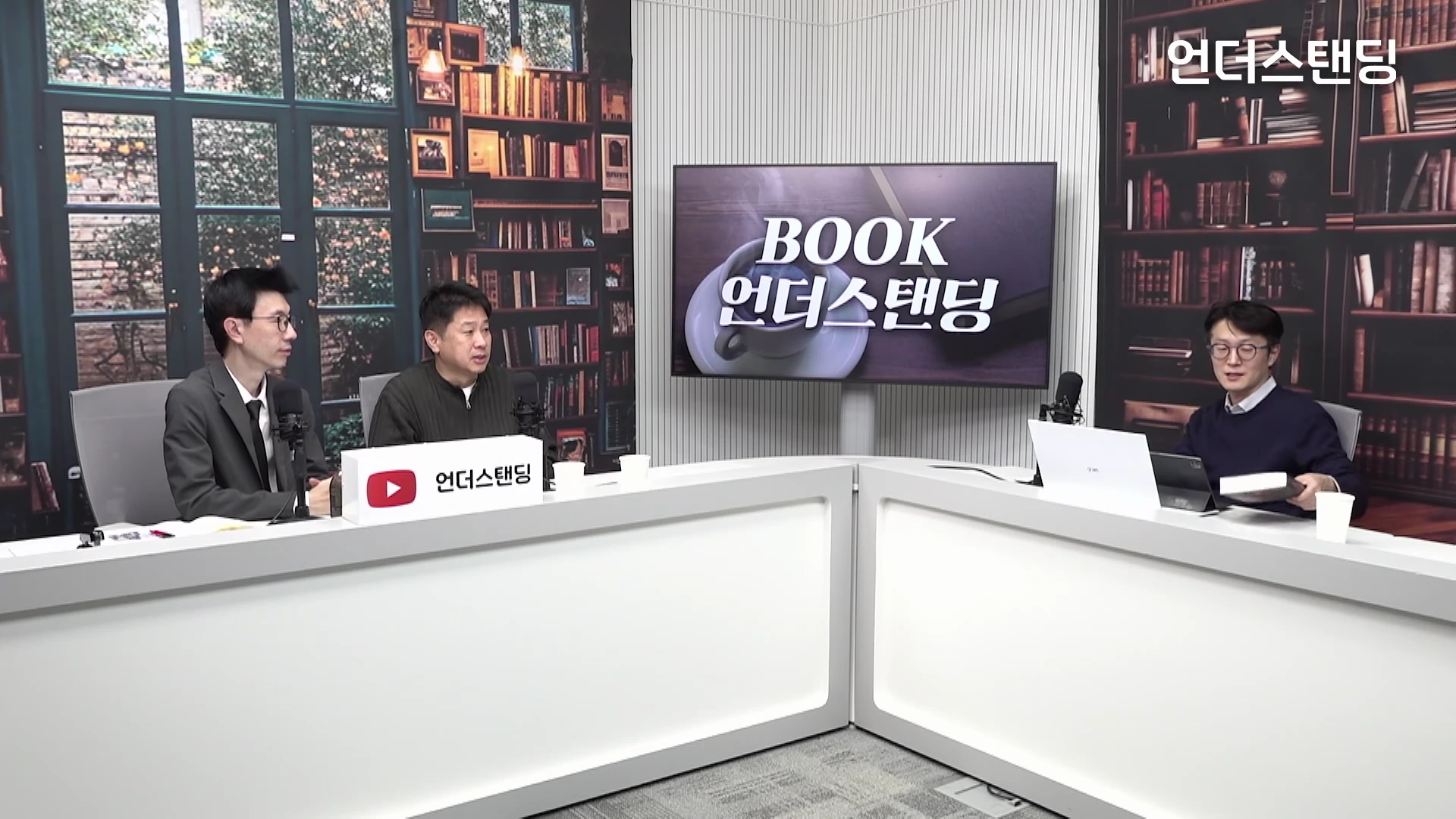 두 명의 남성이 서재 배경의 스튜디오에서 책상을 사이에 두고 마주 앉아 대화하고 있으며, 화면 중앙에는 북 언더스탠딩 로고가 띄워진 모니터가 있습니다.