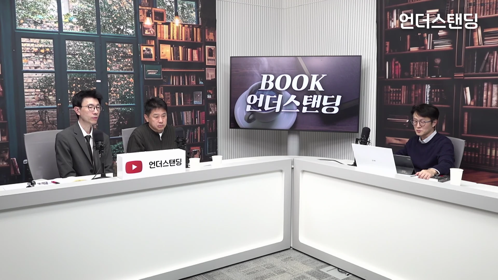 서재 배경의 스튜디오에서 세 명의 남성이 책상에 앉아 마이크를 앞에 두고 대담을 나누고 있는 모습.