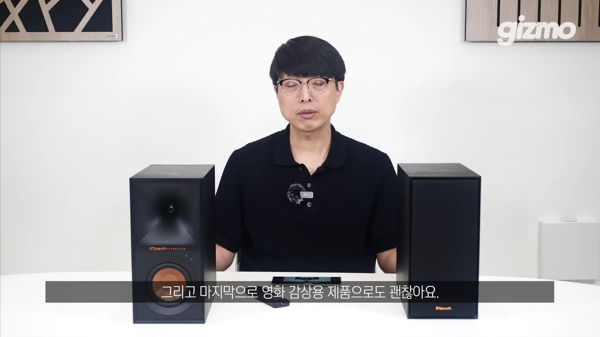안경을 쓴 남성이 테이블 위에 놓인 검은색 북쉘프 스피커 한 쌍을 사이에 두고 앉아 카메라를 바라보며 설명하고 있다.
