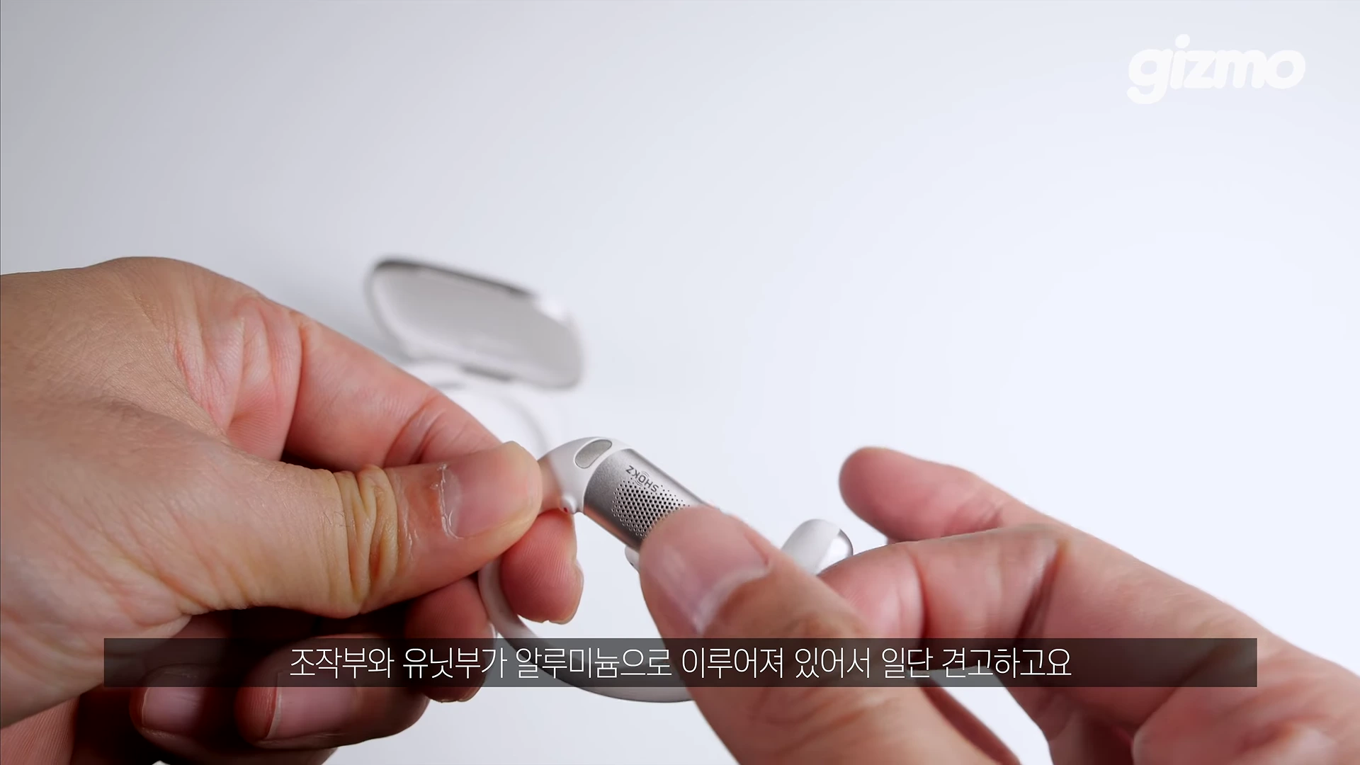 흰색 무선 이어폰 유닛을 양손으로 잡고 살펴보고 있는 모습이며 배경에는 충전 케이스가 놓여 있습니다.