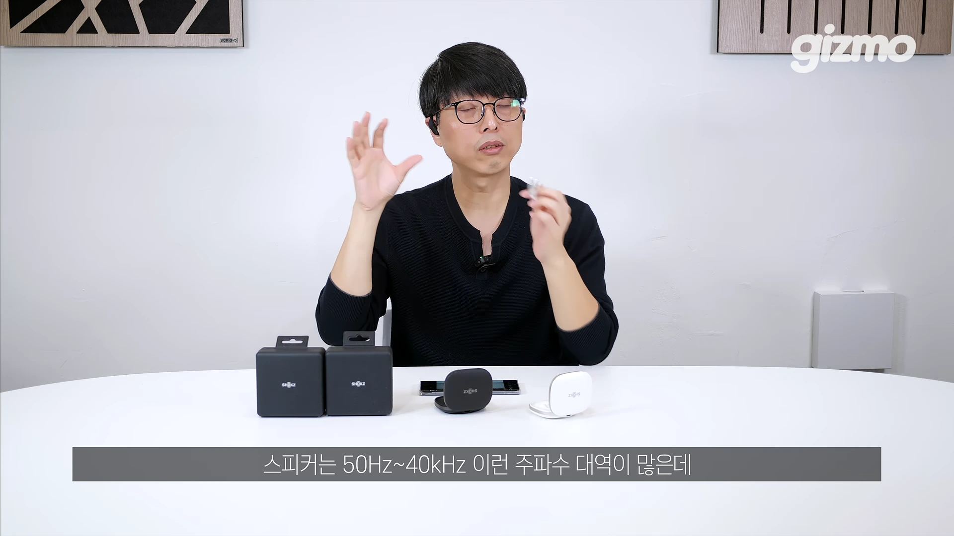 안경을 쓴 남성이 흰색 테이블 위에 놓인 검은색과 흰색 이어폰 케이스들을 앞에 두고 제스처를 하며 설명하고 있는 영상 캡처 화면
