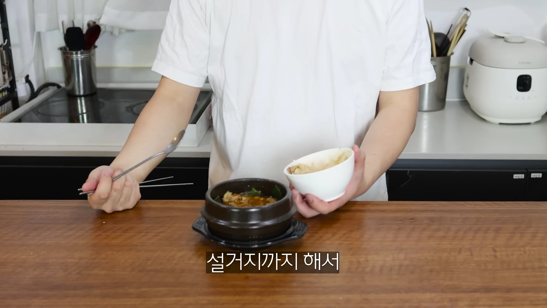 흰색 티셔츠를 입은 사람이 주방 조리대 앞에서 뚝배기와 밥그릇을 들고 있는 모습.