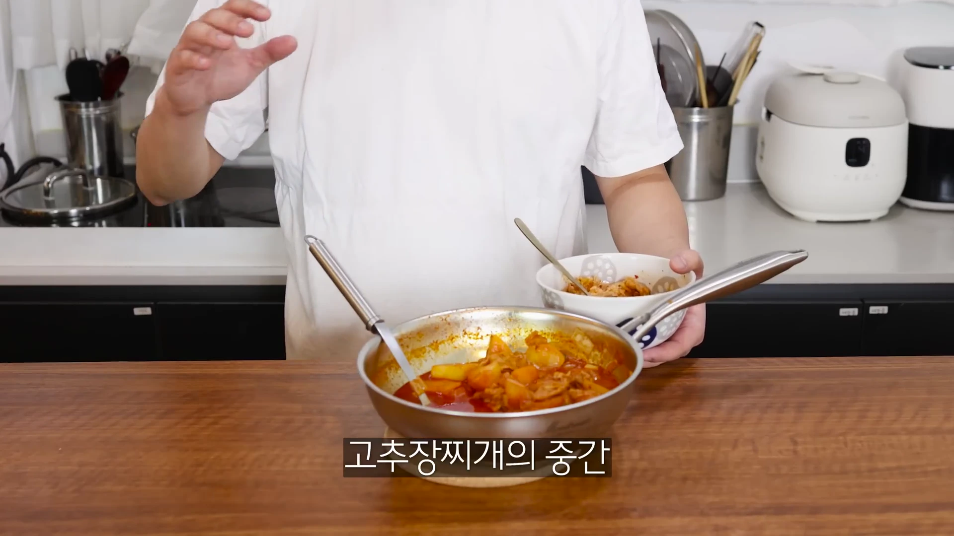 흰 티셔츠를 입은 사람이 프라이팬에 담긴 요리를 앞두고 한 손에 그릇을 든 채 손짓하며 설명하고 있는 모습