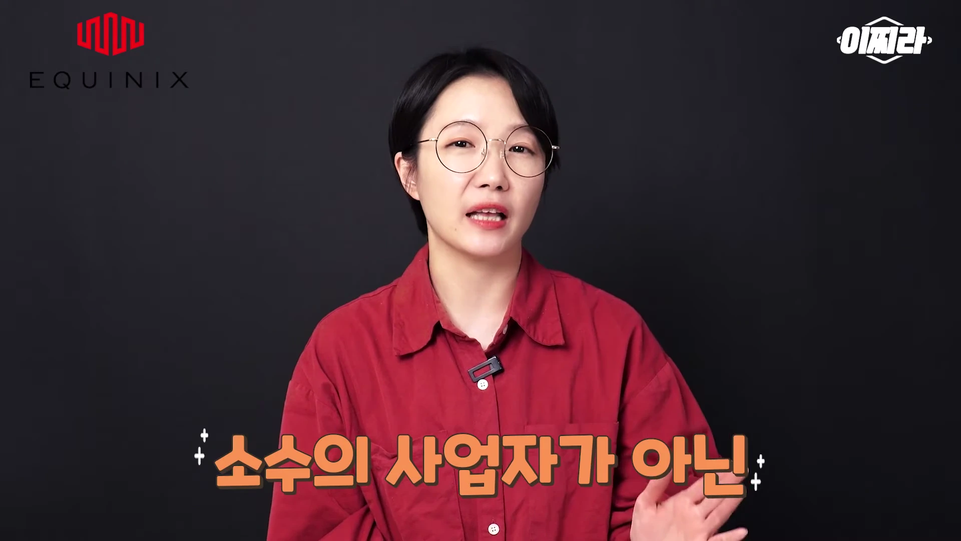 안경을 쓴 여성이 검은색 배경 앞에서 빨간색 셔츠를 입고 이야기를 하고 있으며, 화면 하단에는 '소수의 사업자가 아닌'이라는 자막이 떠 있습니다.