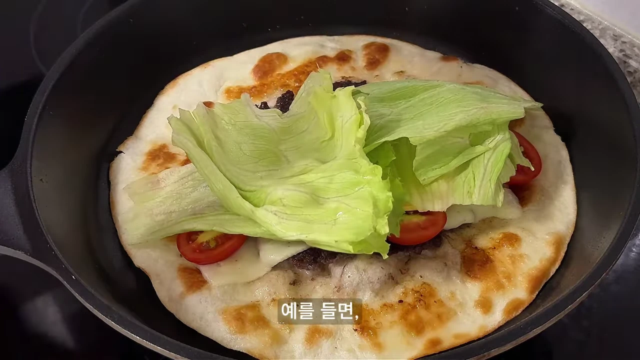 검은색 프라이팬 위에 또띠아와 양상추, 토마토 등 재료가 올라가 있는 모습