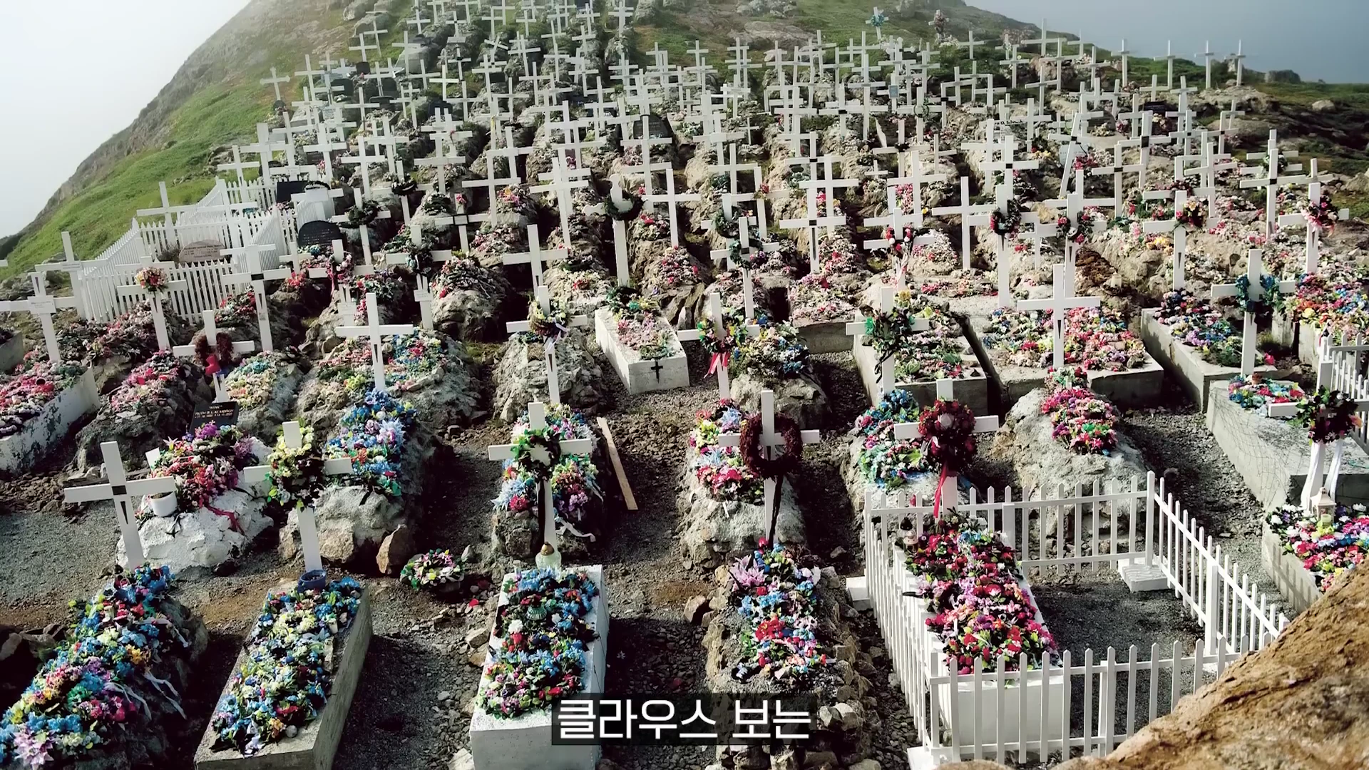 언덕 위에 가득 들어찬 수많은 흰색 십자가 묘비와 그 주위를 덮은 화려한 조화들로 구성된 장례 풍경 사진.